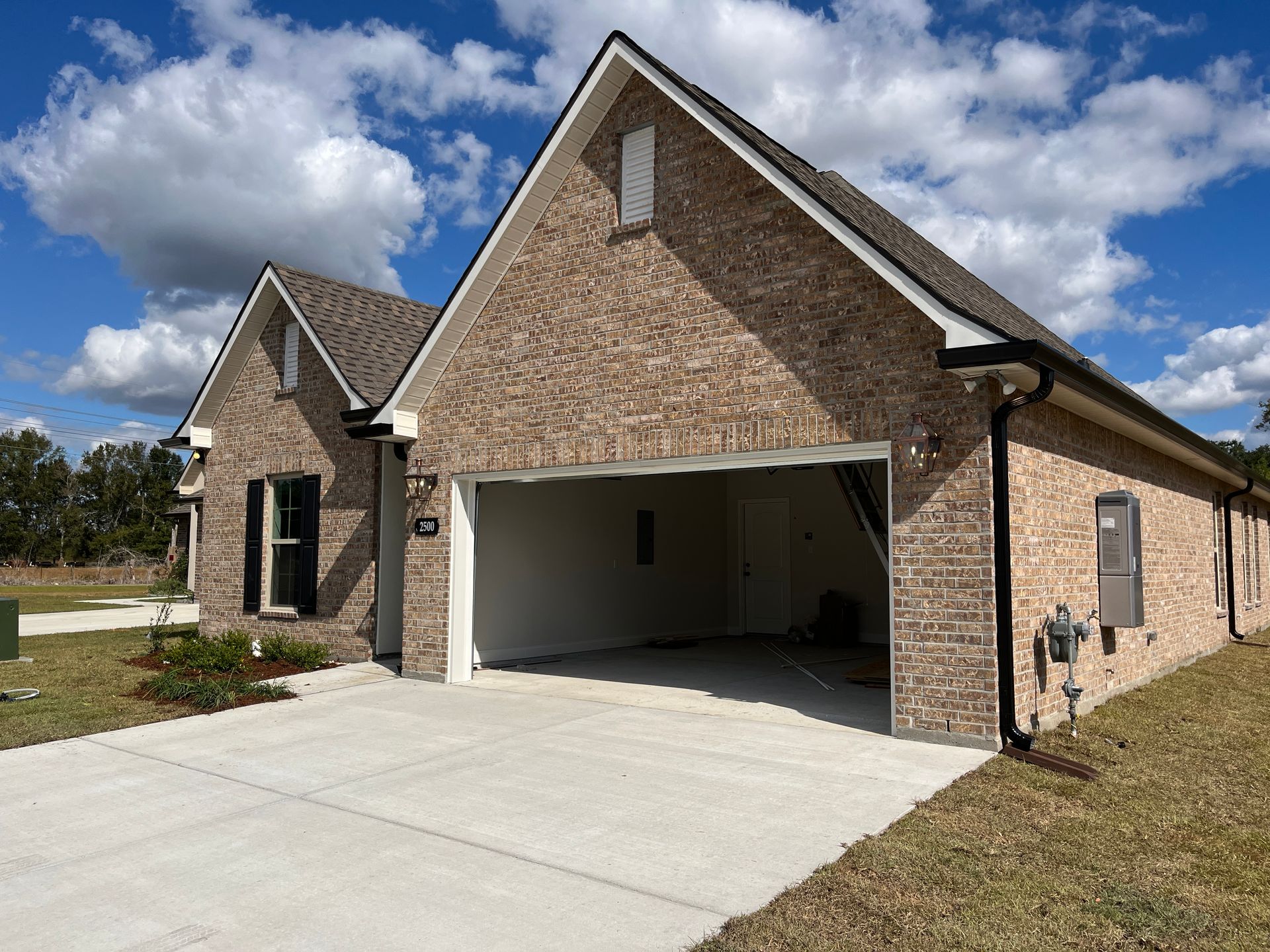 A single-story tan brick house with a dark roof, a white garage interior, and black shutters under a blue, cloudy sky.