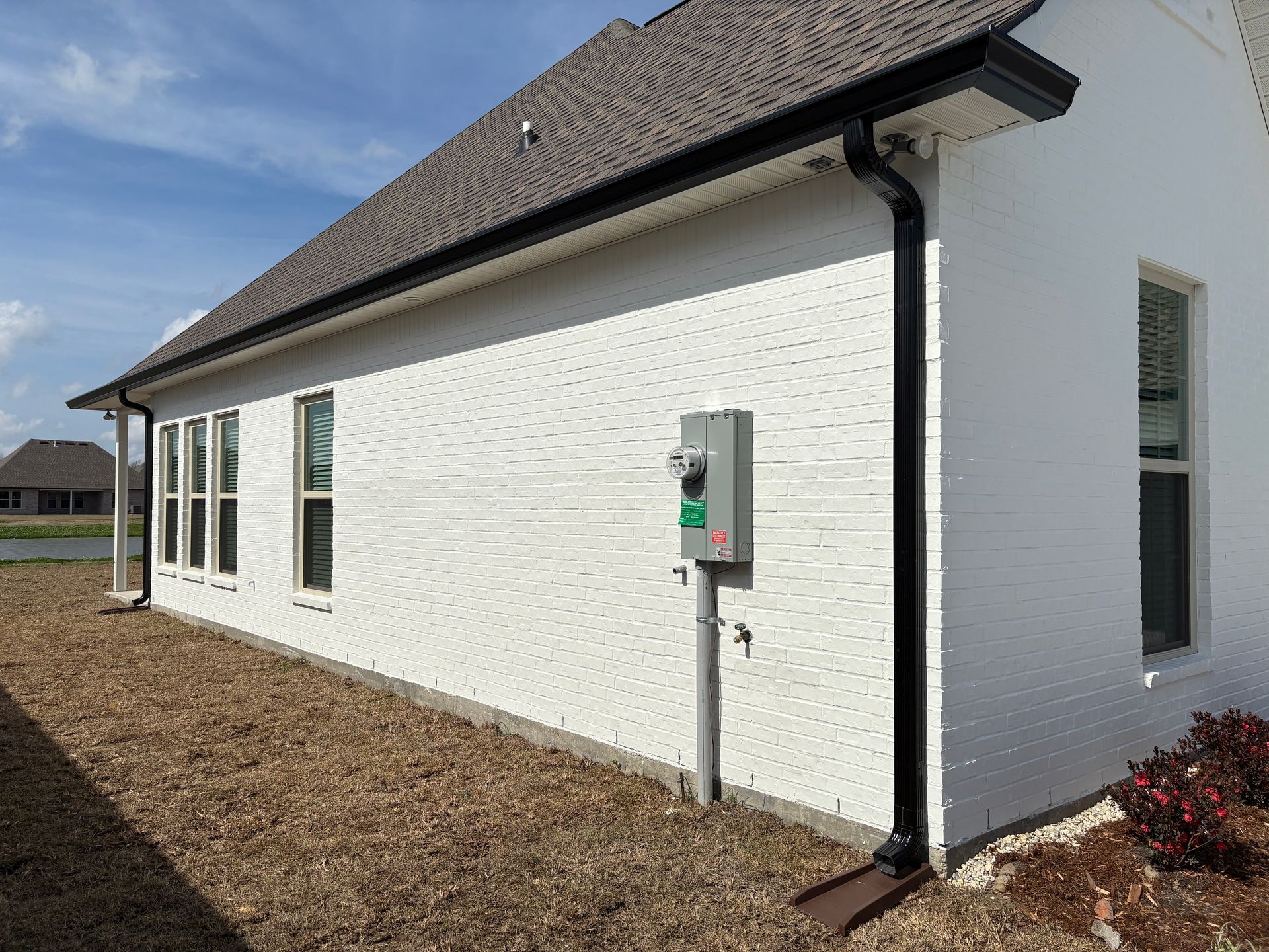 A side view of a white brick house with black gutters and a gray electrical meter box mounted on the exterior wall.