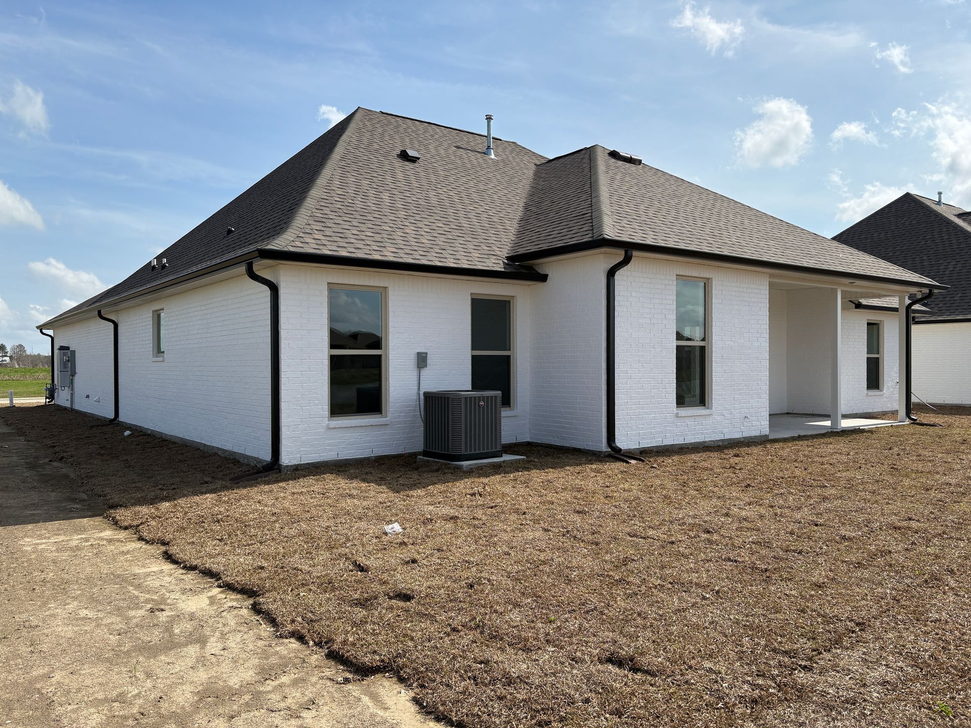 Rear view of a new white brick house with a dark shingle roof and black gutters on an unfinished dirt lot.