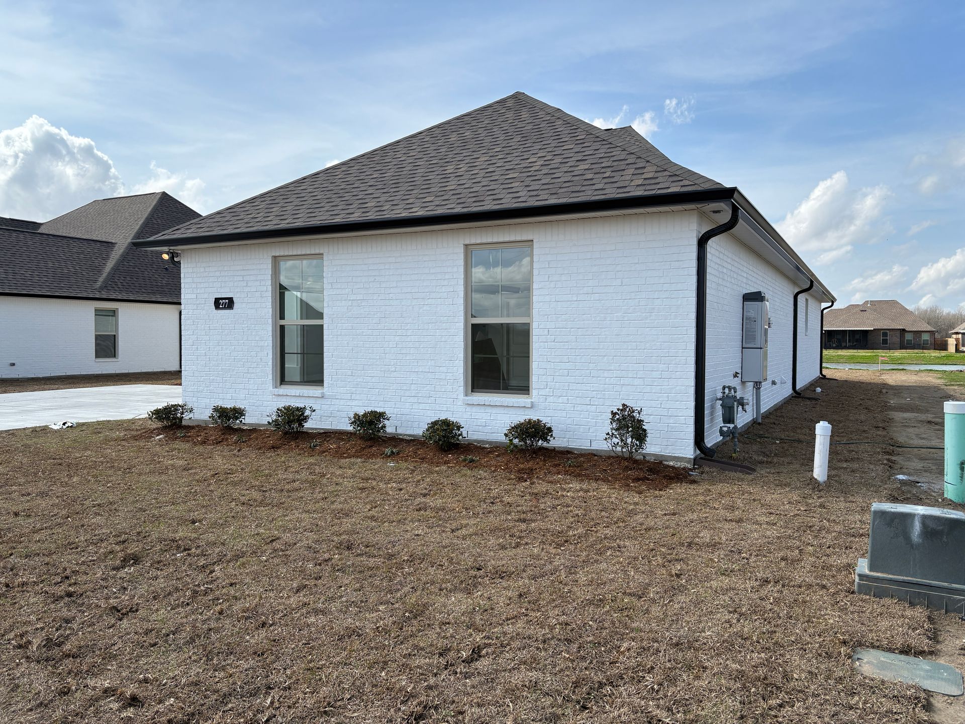 A single-story house with a white textured exterior, two windows, a dark roof, and a wood-mulched yard under a blue sky.