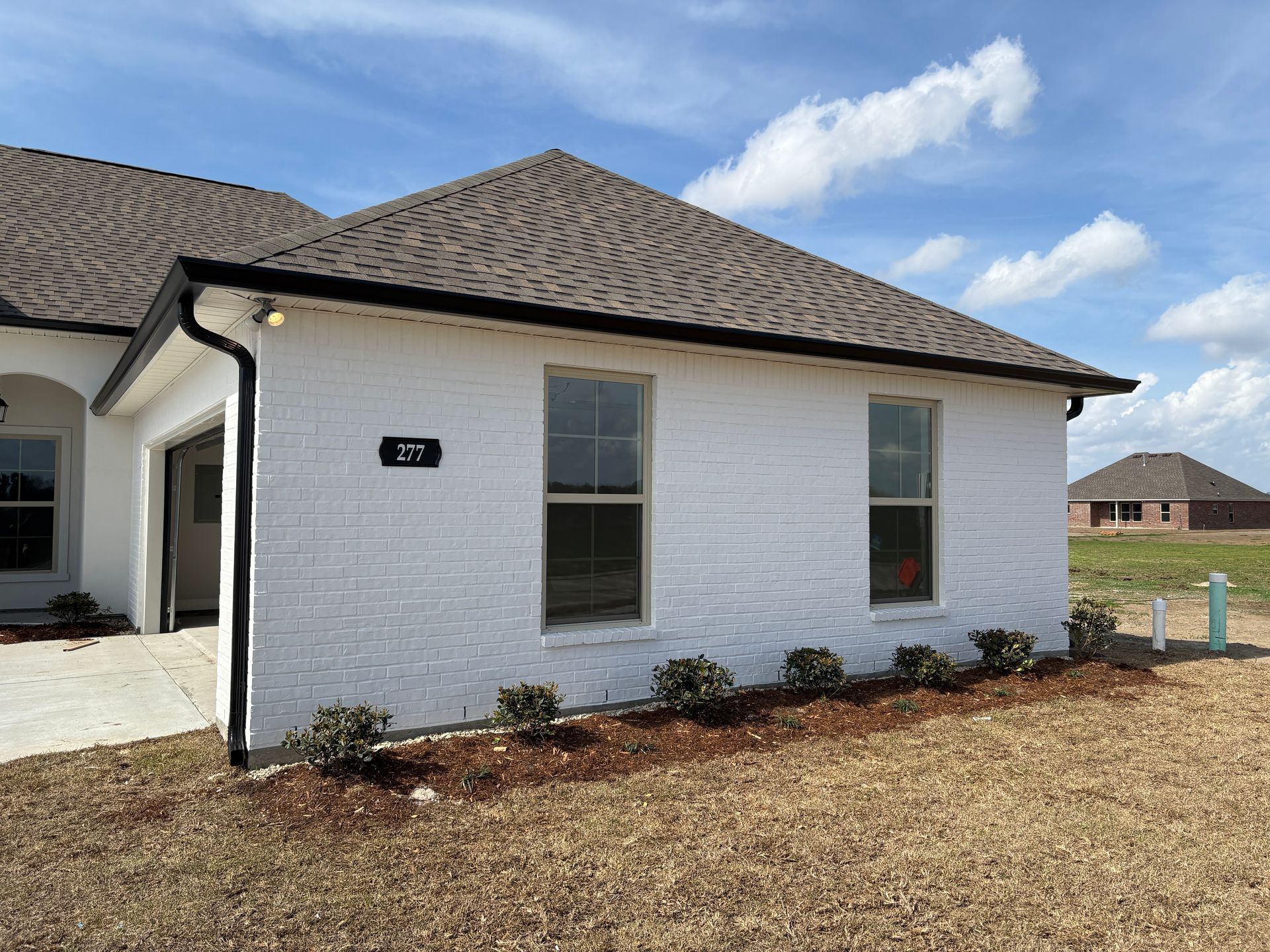 A white brick house with a dark roof and black gutters sits under a blue sky with scattered clouds.