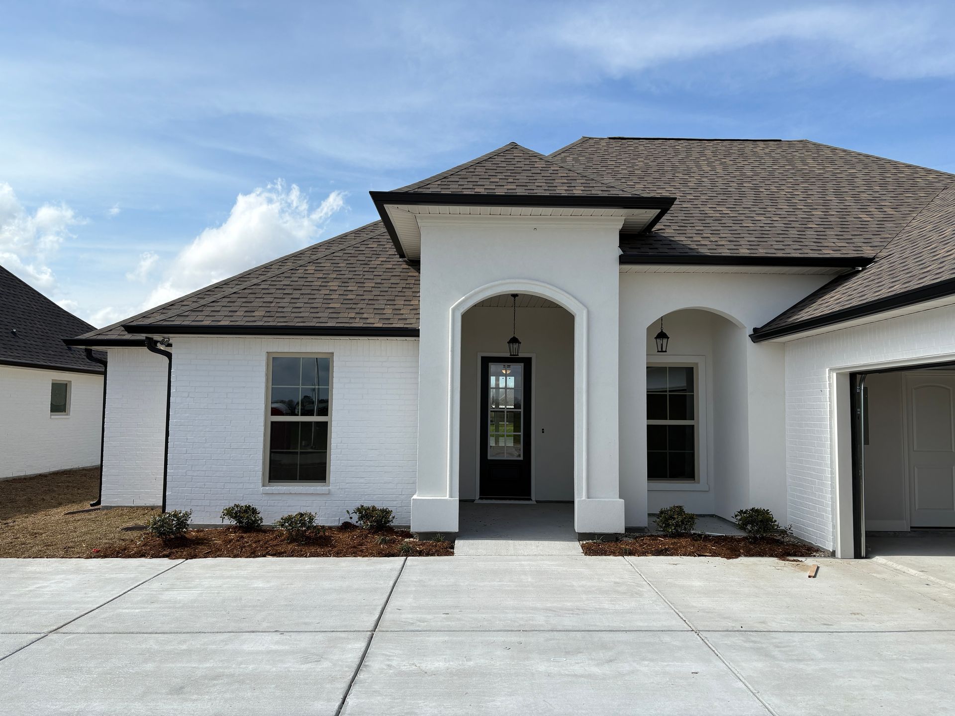 A white brick house with a dark roof and a concrete driveway under a blue, cloudy sky.