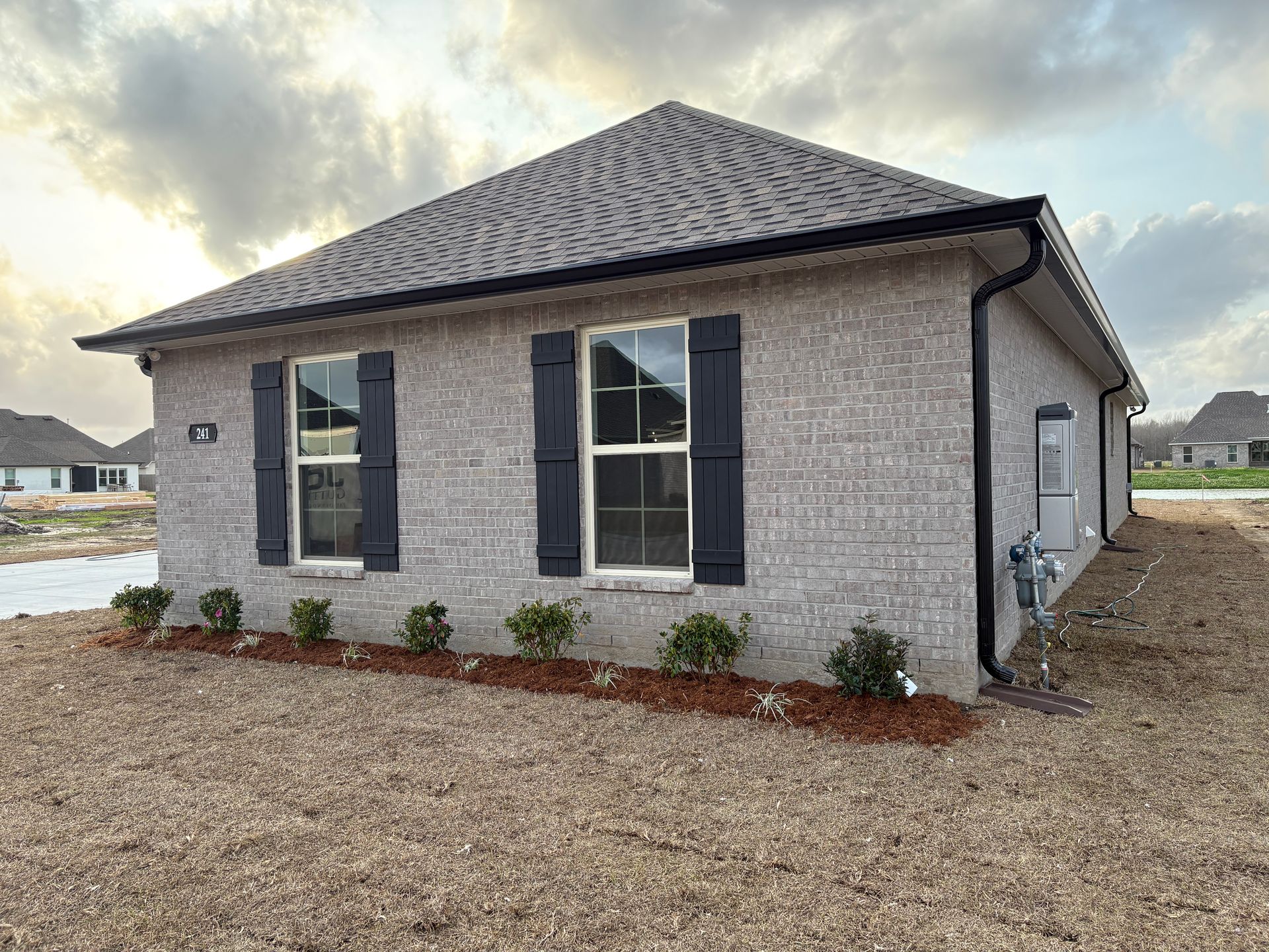 A single-story gray brick house with black shutters on two windows, surrounded by landscaping mulch on a gravel lot.