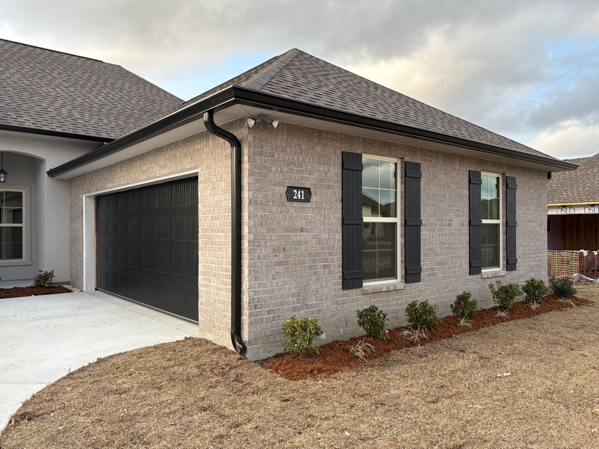 A light-colored brick residential house with a dark garage door, roof shingles, and shutters under an overcast sky.