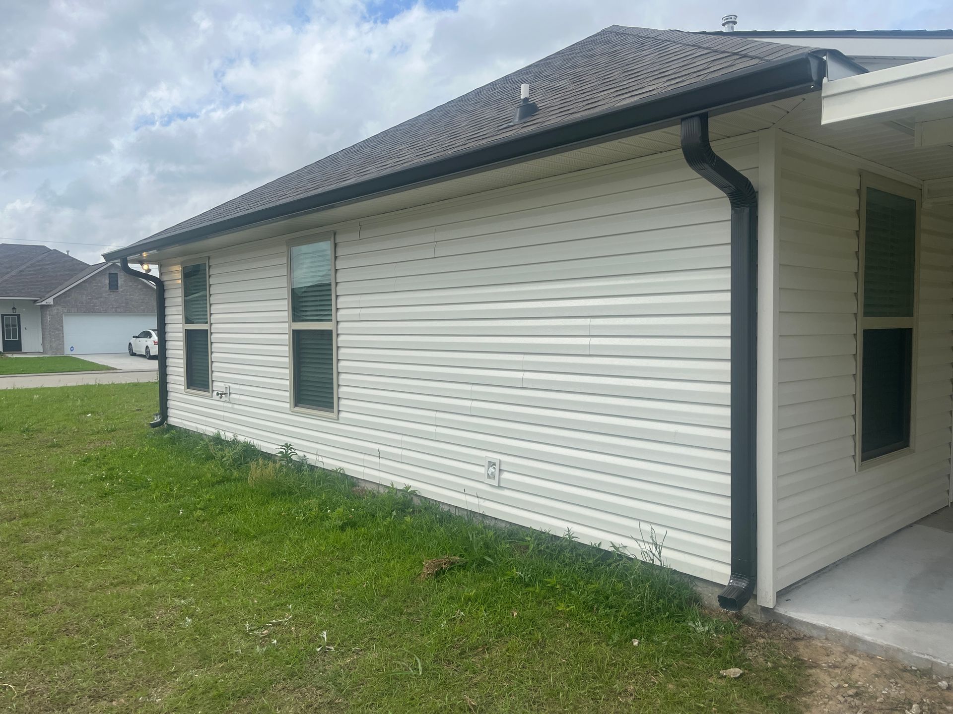 A side view of a house with beige vinyl siding, dark gutters, and two windows, set in a yard under a cloudy sky.