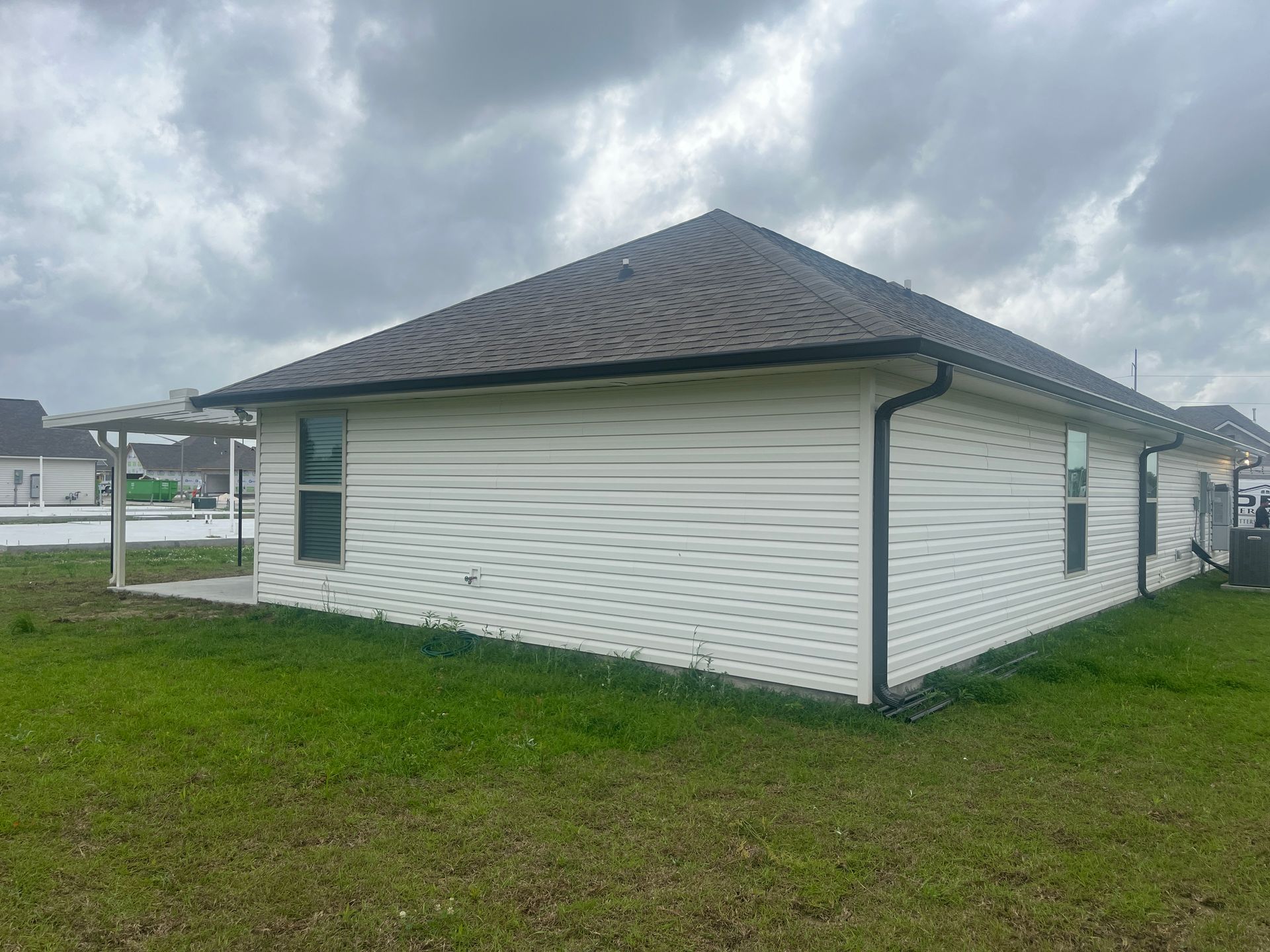 A beige, single-story house with a dark roof and white trim seen from the side, situated on a grassy lot under cloudy skies.