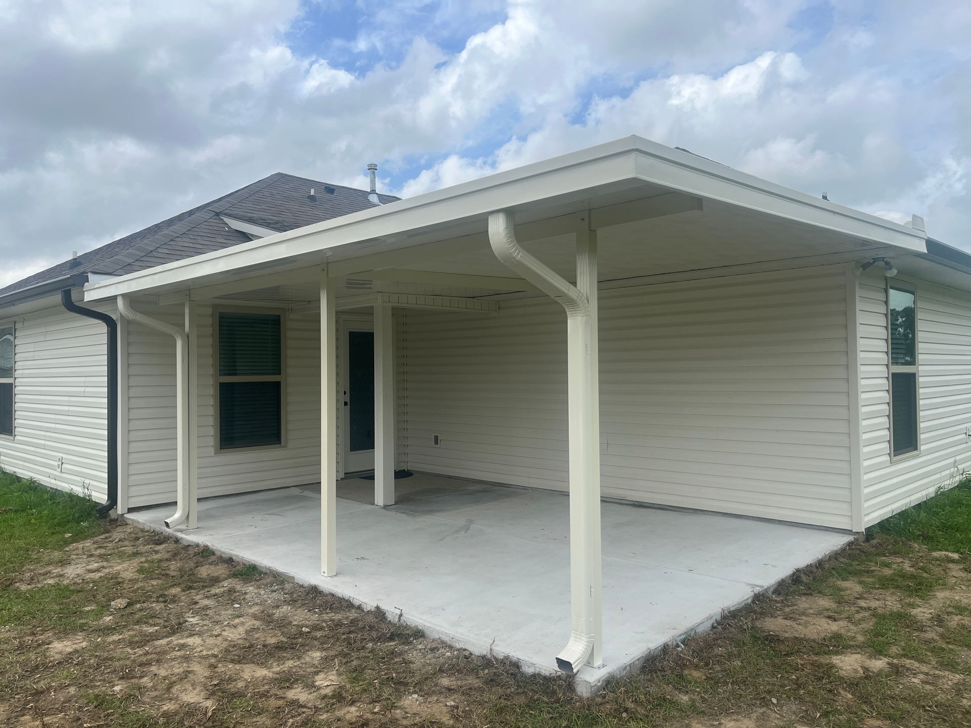 A covered concrete patio attached to the back of a beige, vinyl-sided house under a cloudy sky.