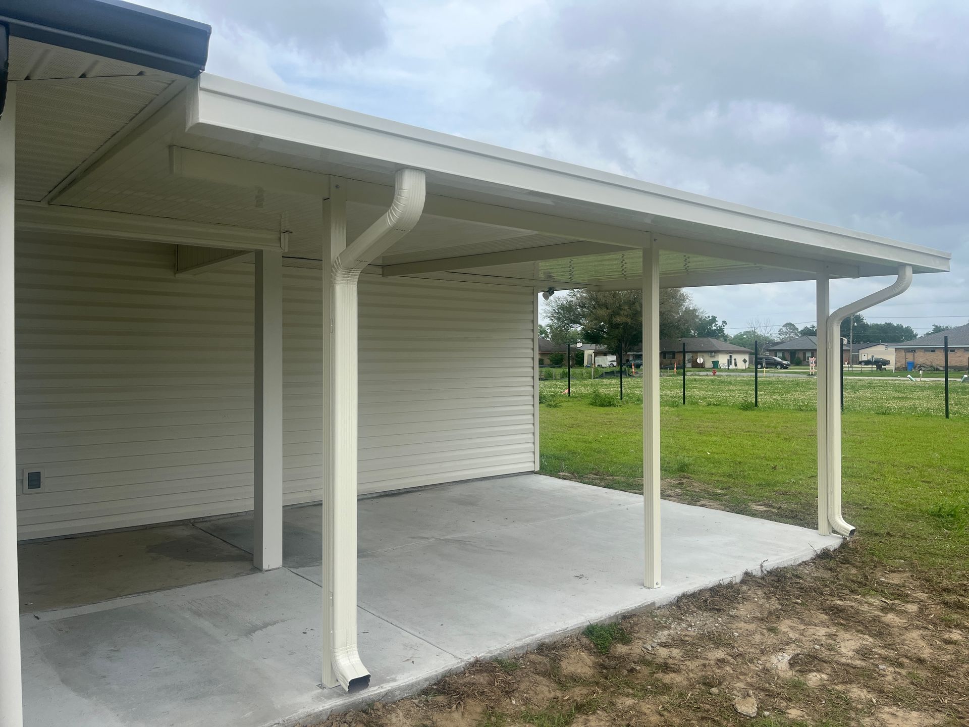 A covered light-colored patio attached to a house with beige siding, overlooking a green grassy field under a cloudy sky.