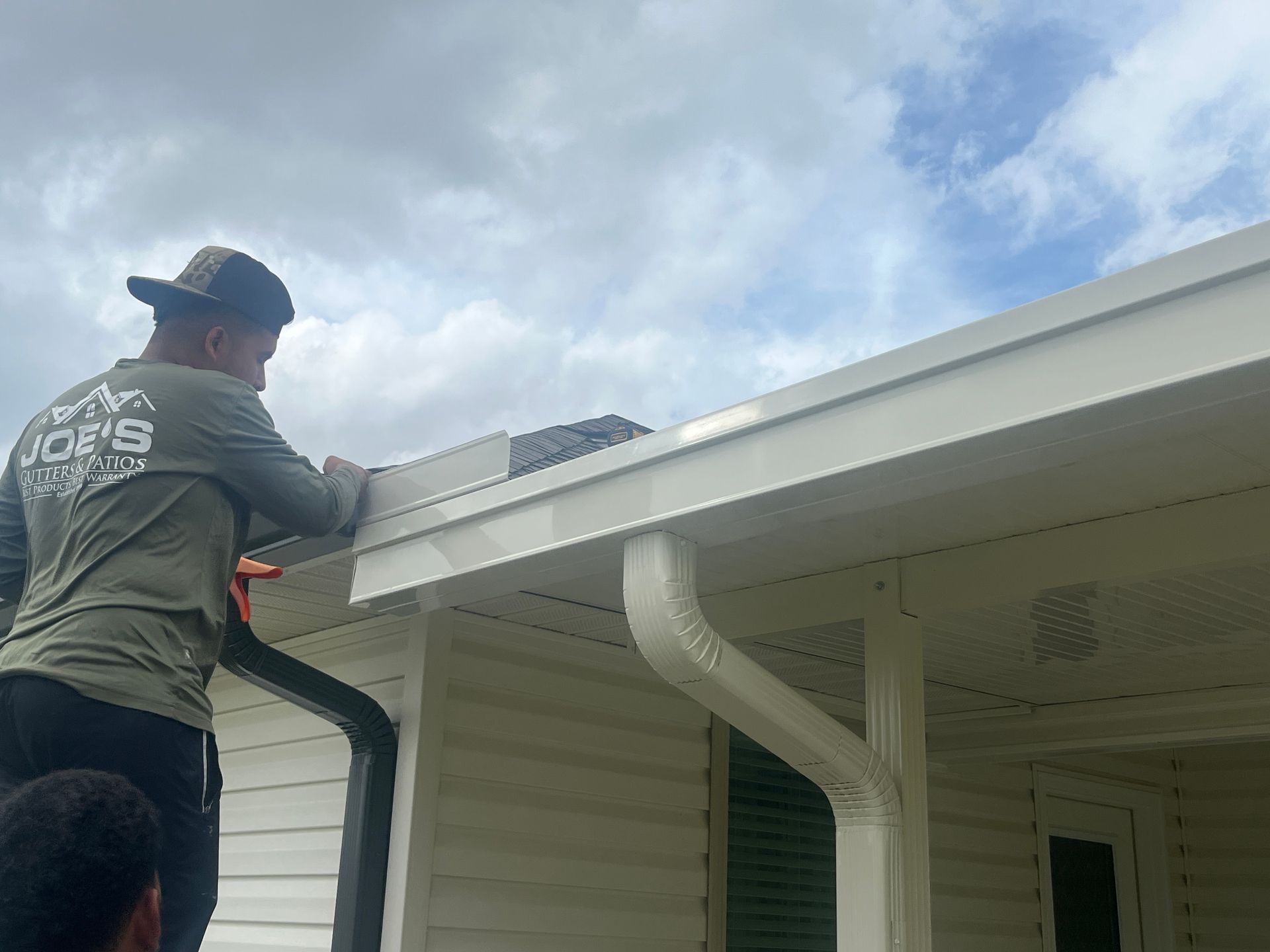 A worker in a gray shirt installs a white gutter section on the edge of a house roof under a cloudy sky.