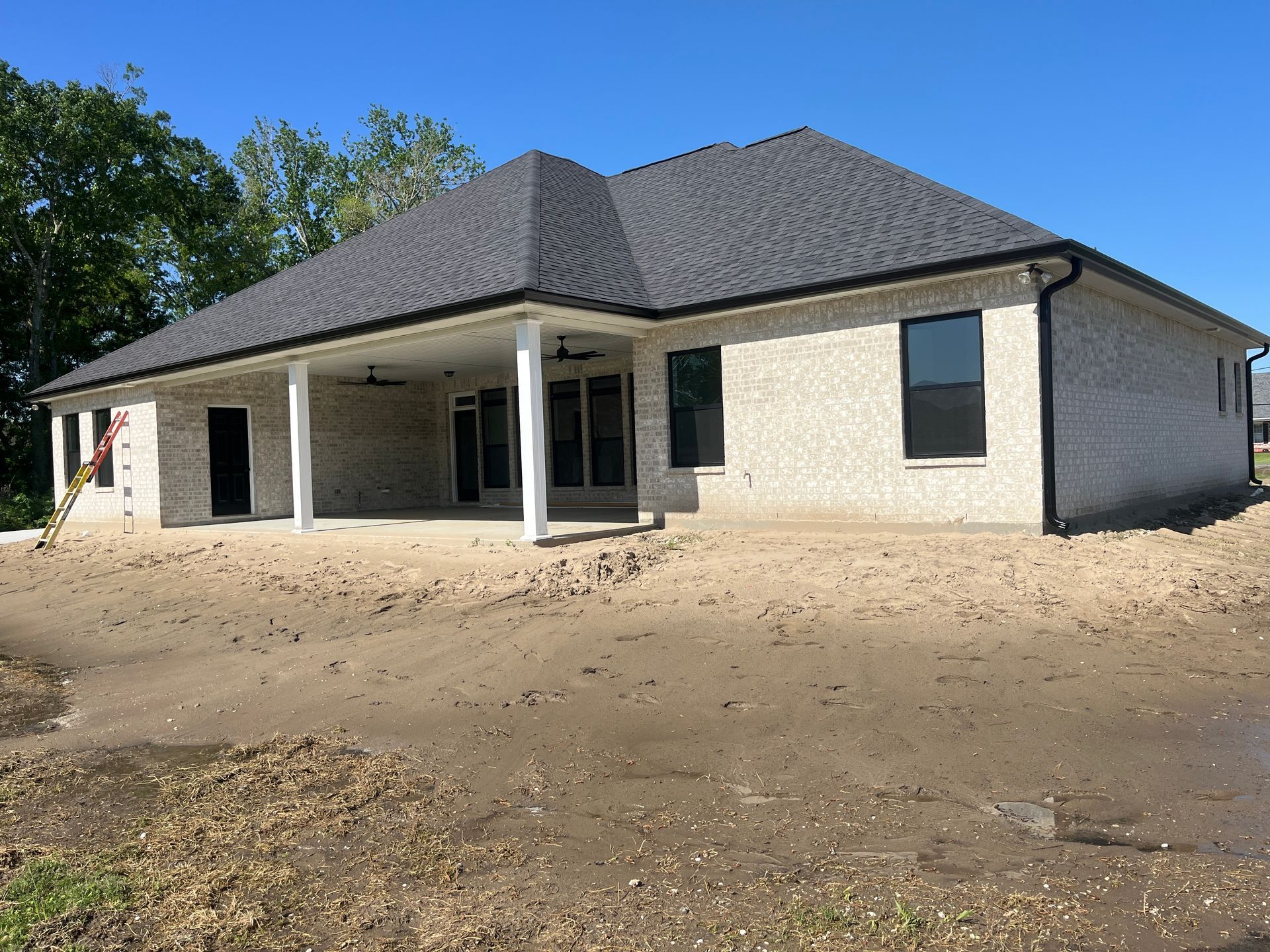 A single-story, light-colored brick house with a dark roof and a covered back patio under a clear blue sky.