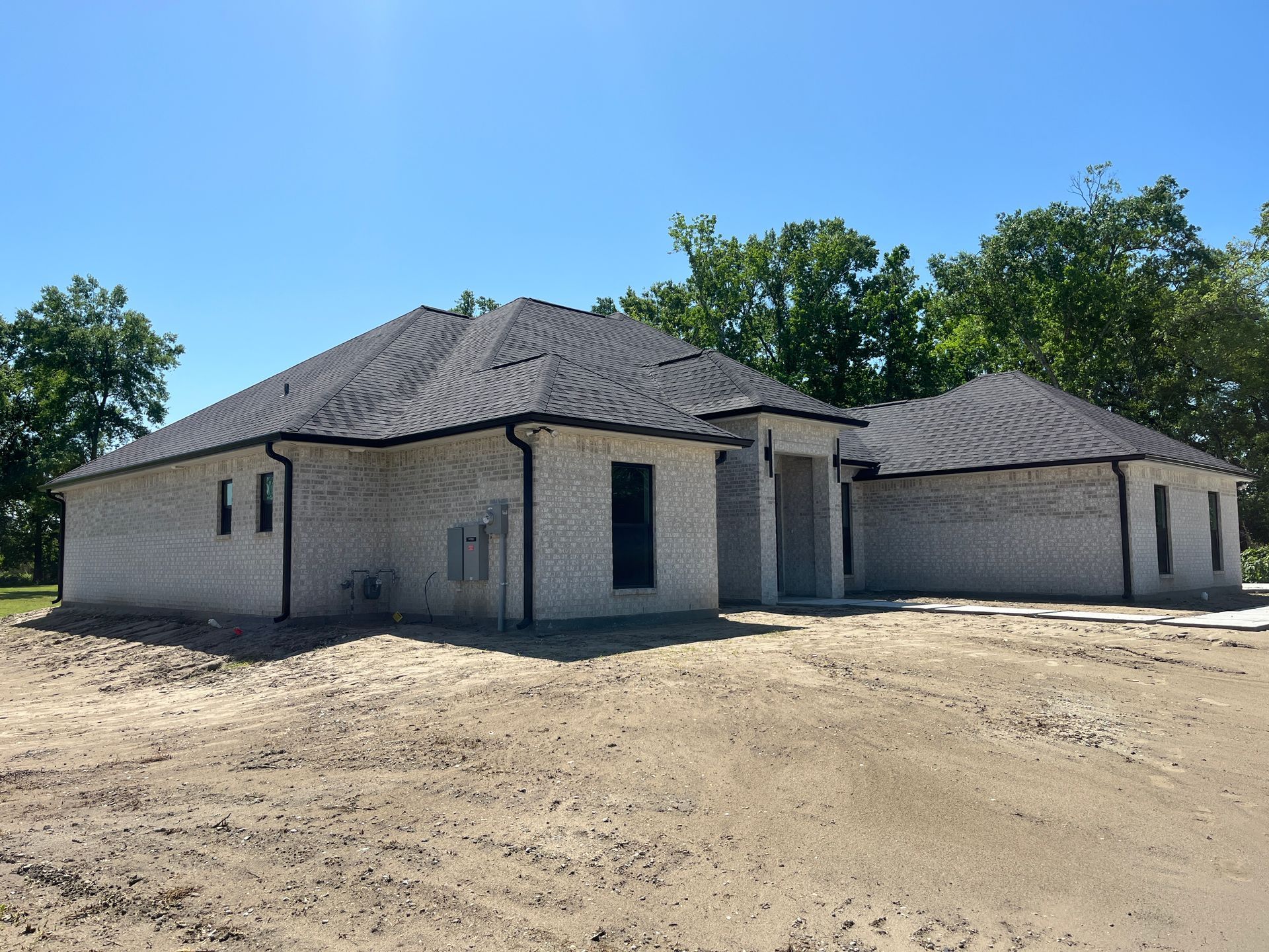 A single-story, light-colored brick house with a dark roof stands on an unlandscaped dirt lot under a clear blue sky.
