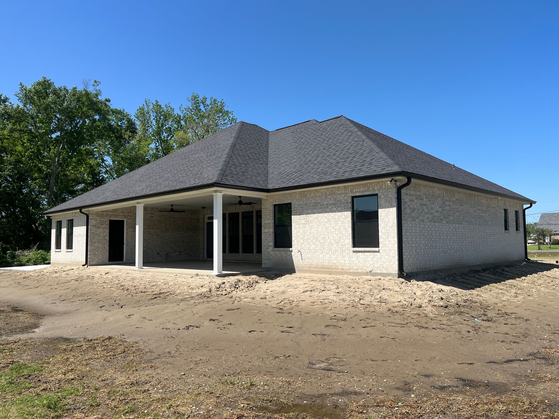 A new one-story, light-colored brick house with a dark roof and a covered back patio, set on a bare dirt lot.