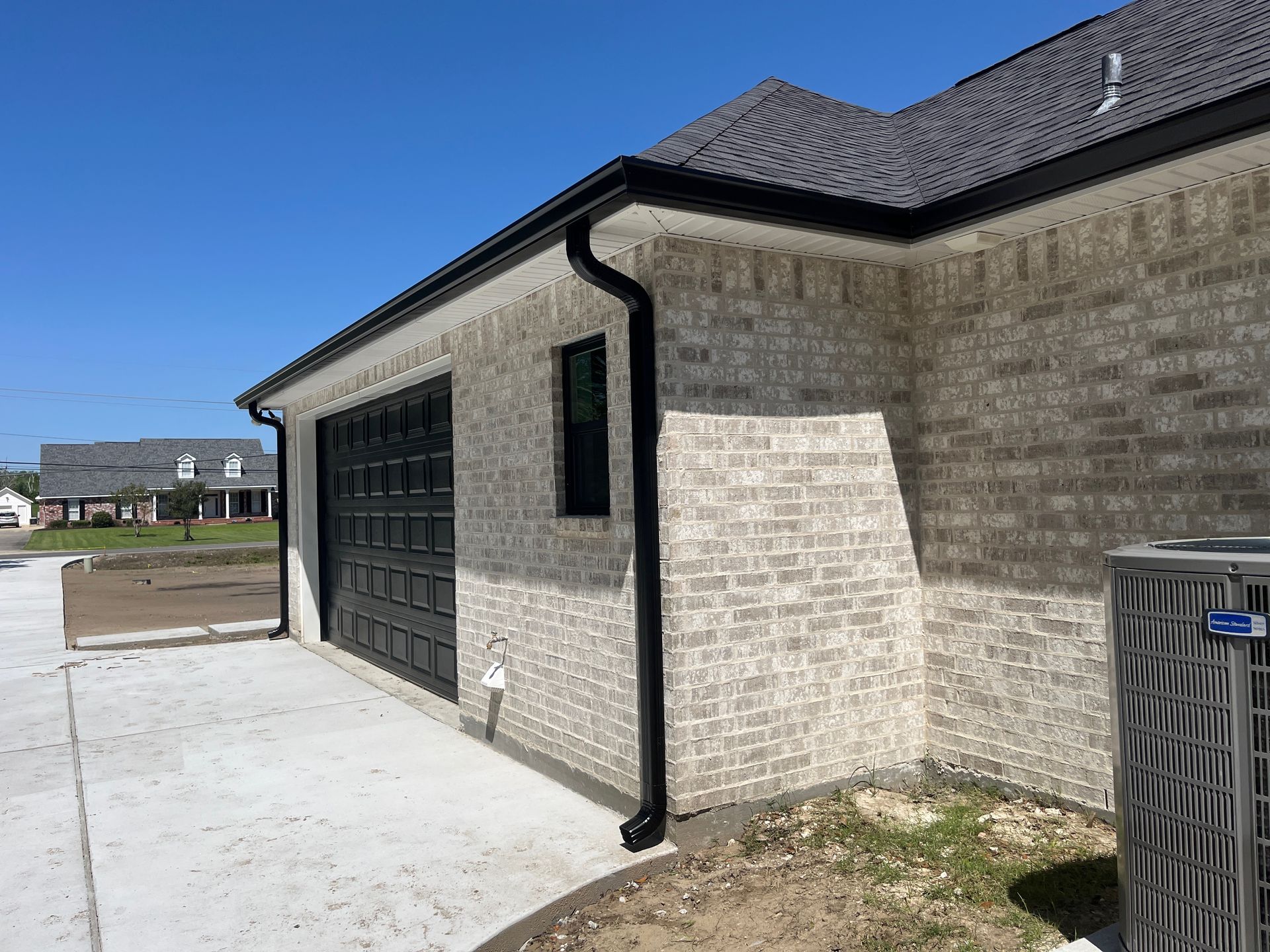 A light-colored brick garage exterior featuring a black door, black gutters, and an adjacent HVAC unit under a blue sky.
