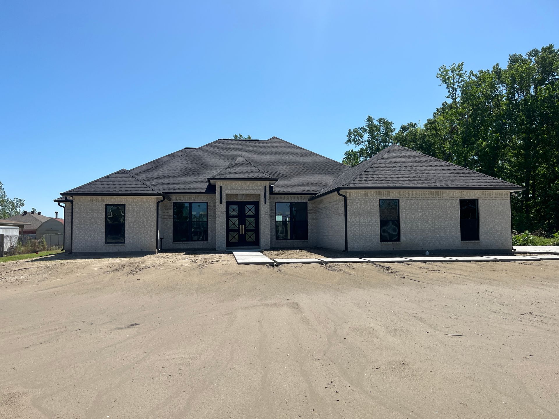 A newly built, one-story brick house with a dark tiled roof and large windows, situated on an unfinished dirt lot.