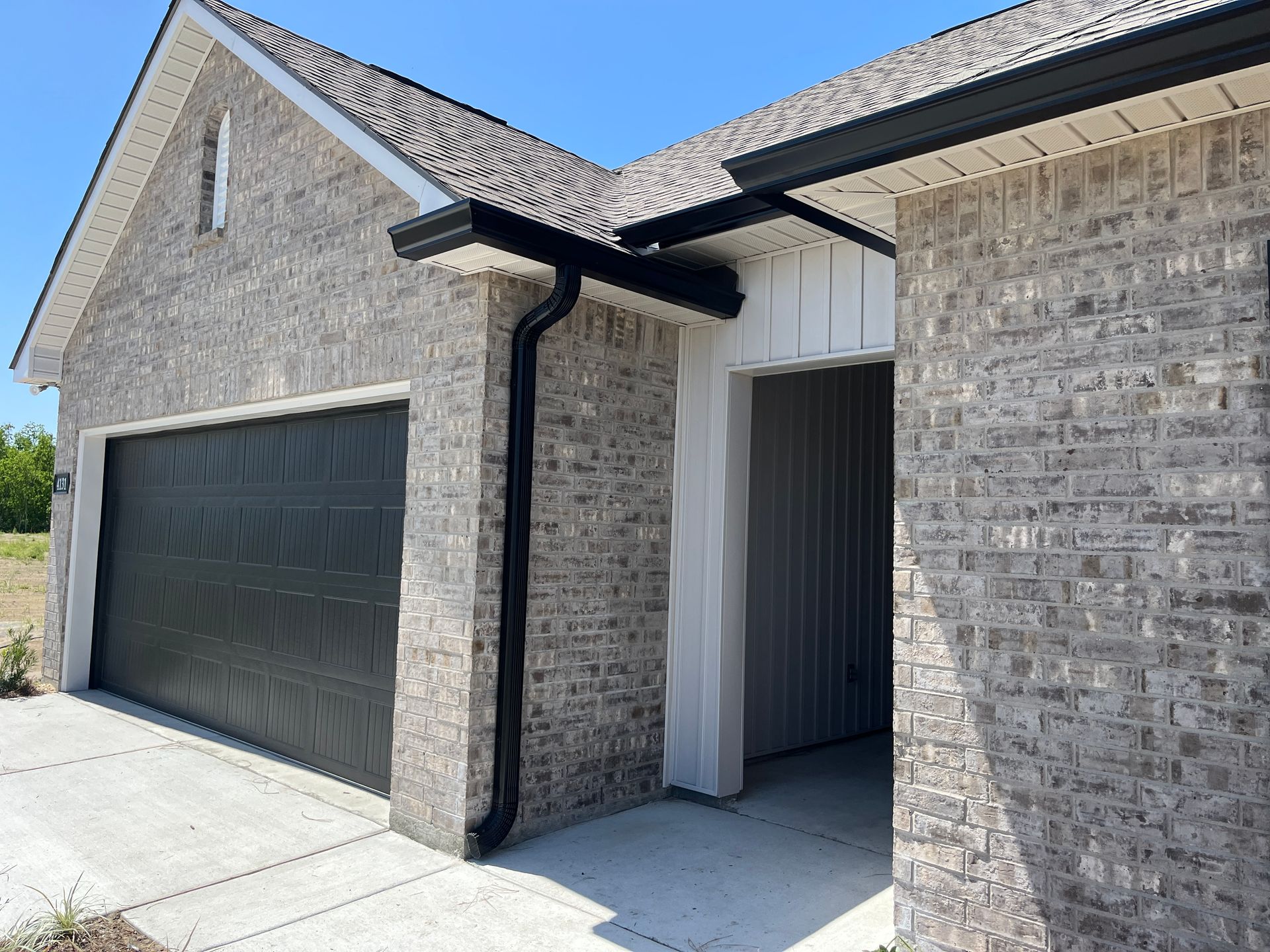 A gray brick house exterior with a black garage door, matching black gutters, and a recessed entryway under a blue sky.