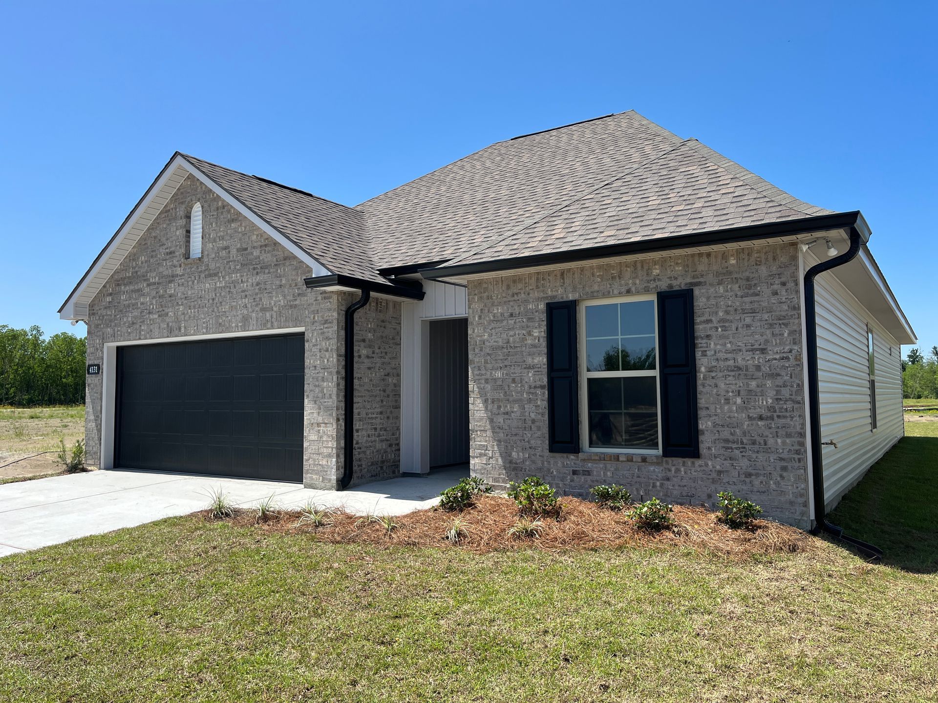 A single-story house with gray brick, a dark gray garage door, black shutters, and a shingled roof under a clear blue sky.