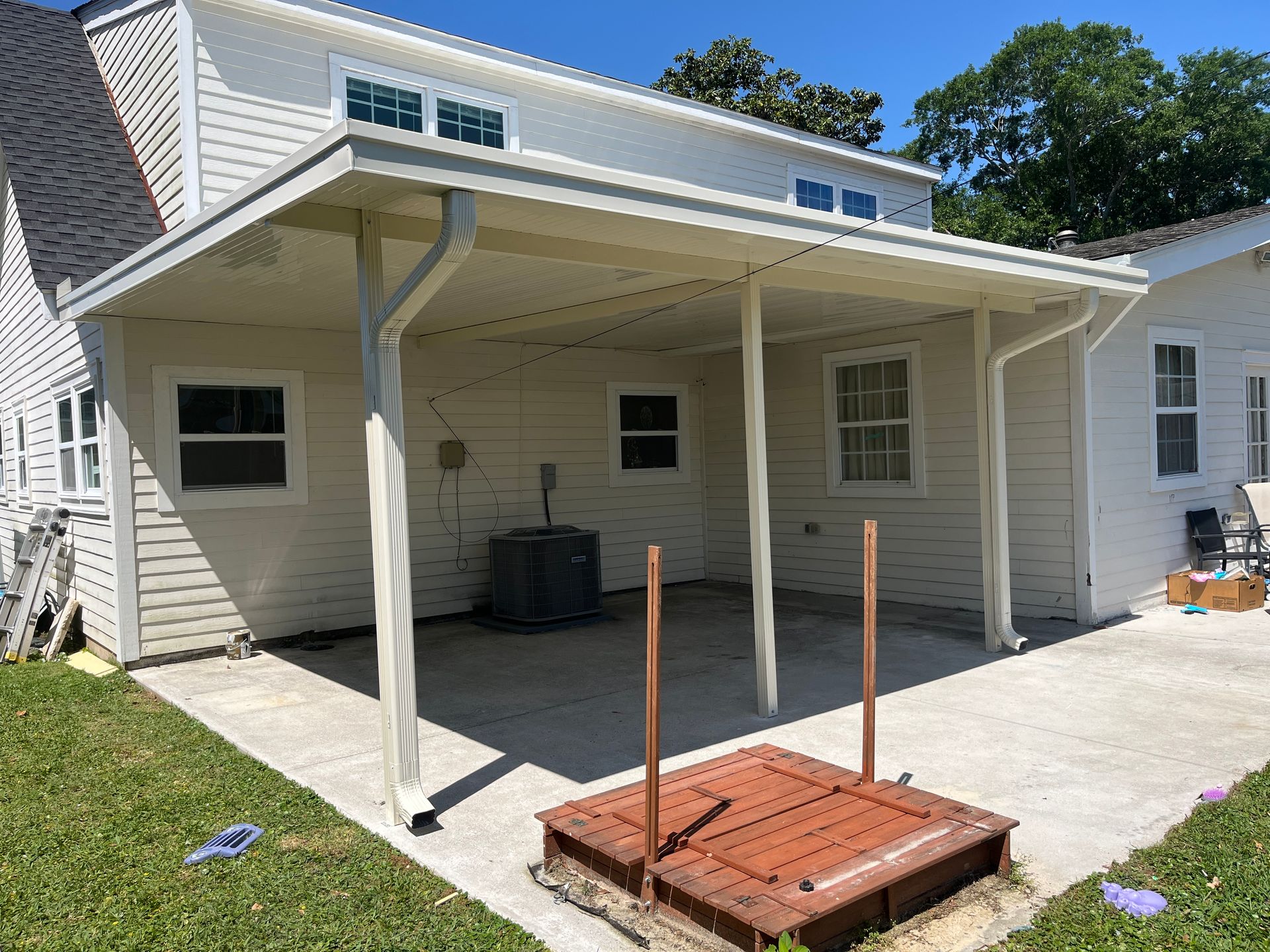 A white house with a covered concrete patio and a raised, wooden rectangular platform in the foreground.