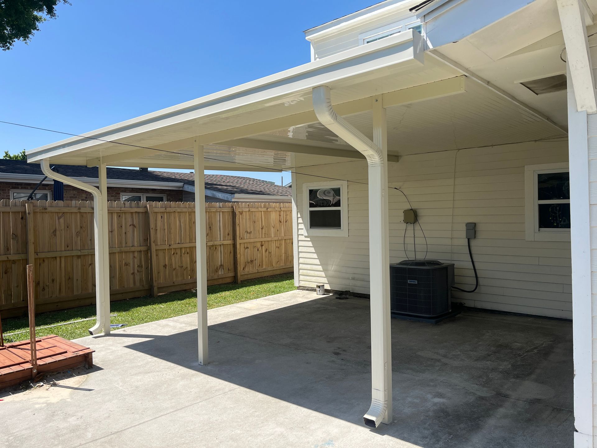A cream-colored metal patio cover attached to a house, with white support beams, gutters, and a concrete floor.
