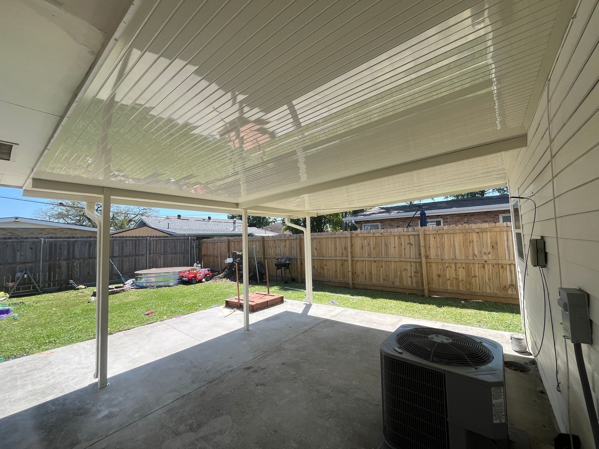 A concrete backyard patio featuring a white metal patio cover, an AC unit against the wall, and a wooden privacy fence.