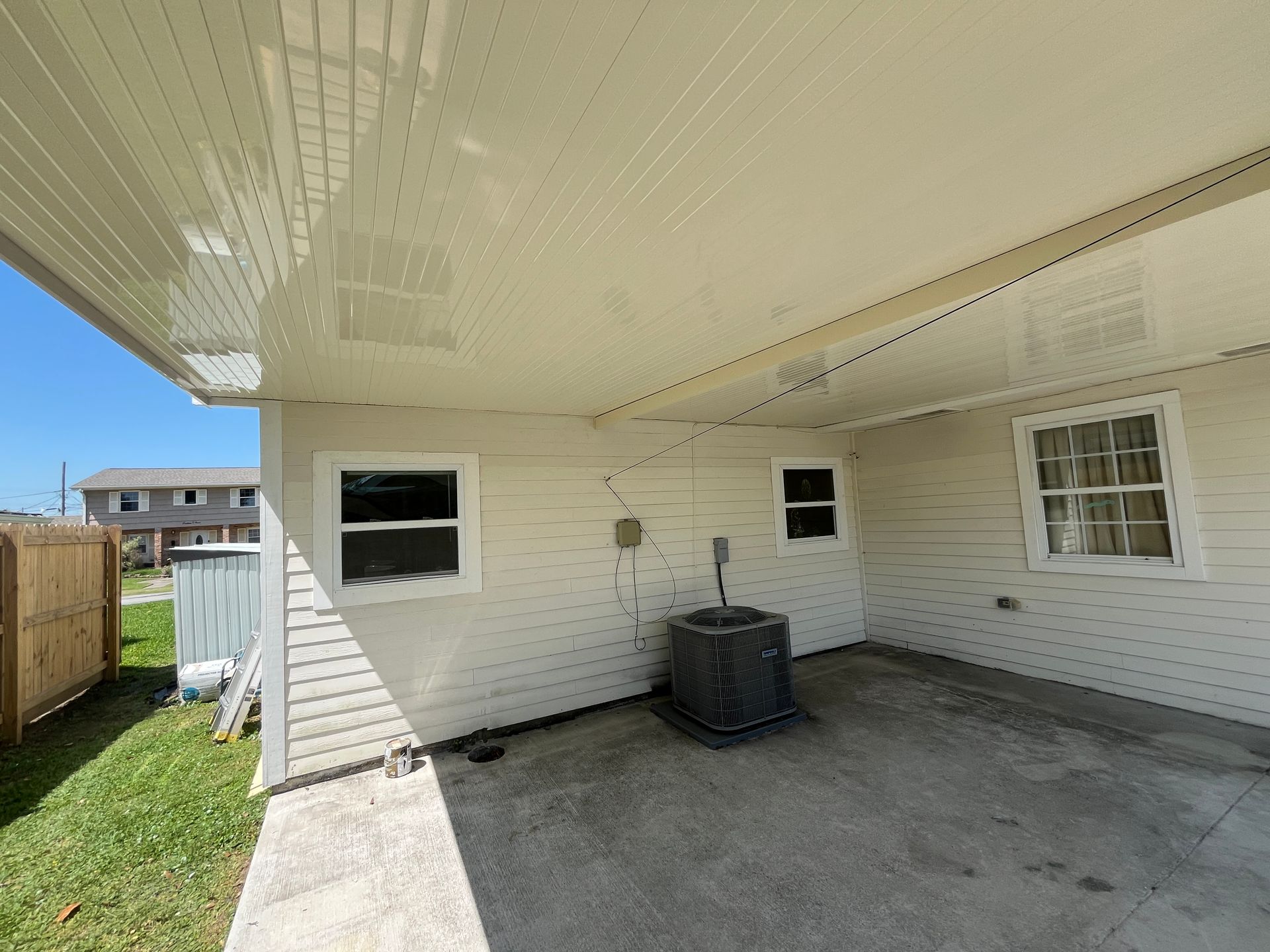 A view of a patio area with light-colored siding, two windows, and an outdoor HVAC unit under a covered porch roof.