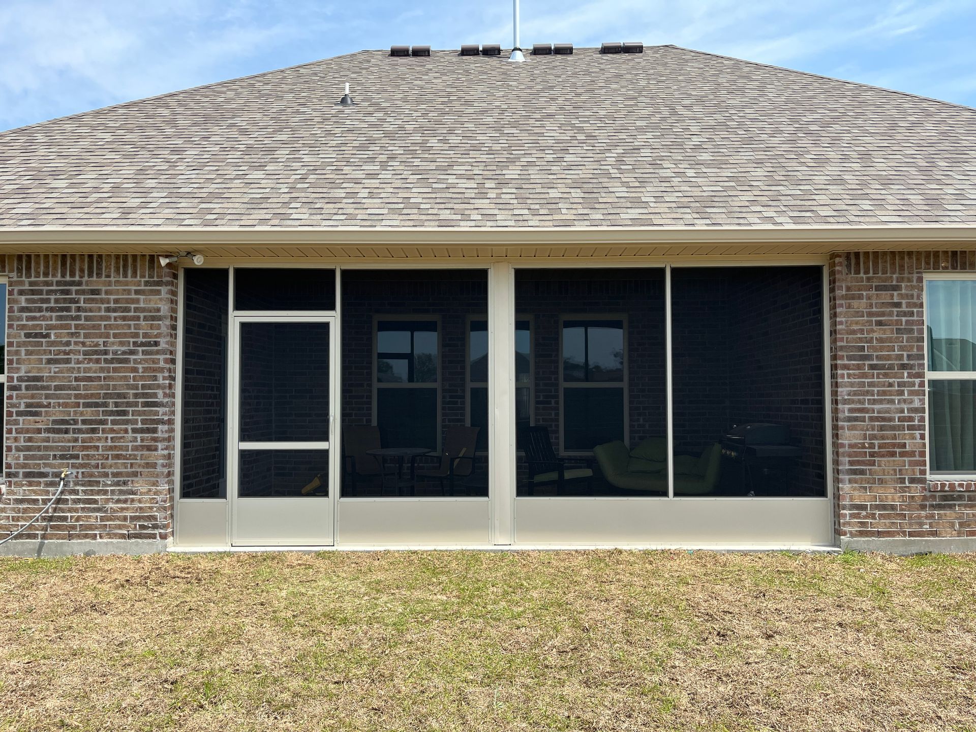 A brick house exterior featuring a screened-in porch with a white door and windows, viewed from a dry grass backyard.