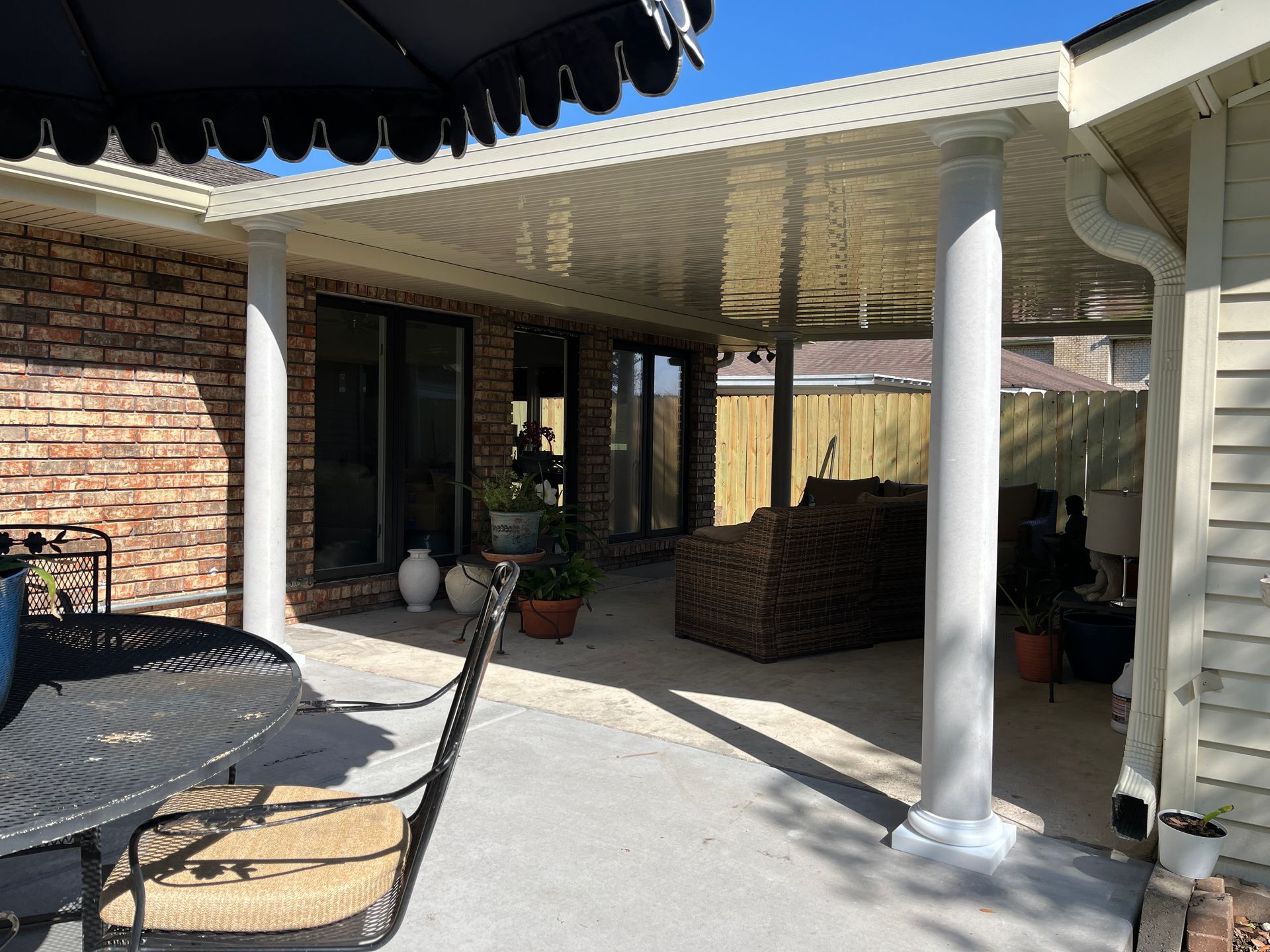 A covered patio with a white ceiling and columns, featuring outdoor furniture on a concrete floor next to a brick house.