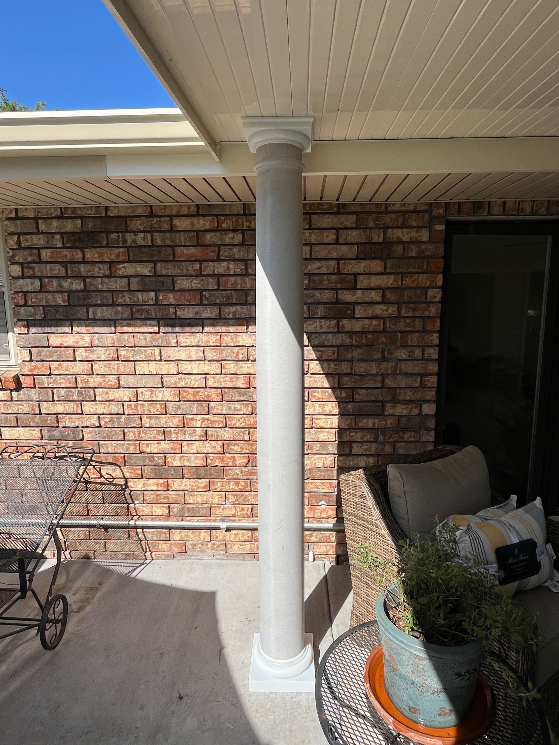 A gray cylindrical patio support pillar stands against a textured red-and-brown brick wall on a concrete porch.