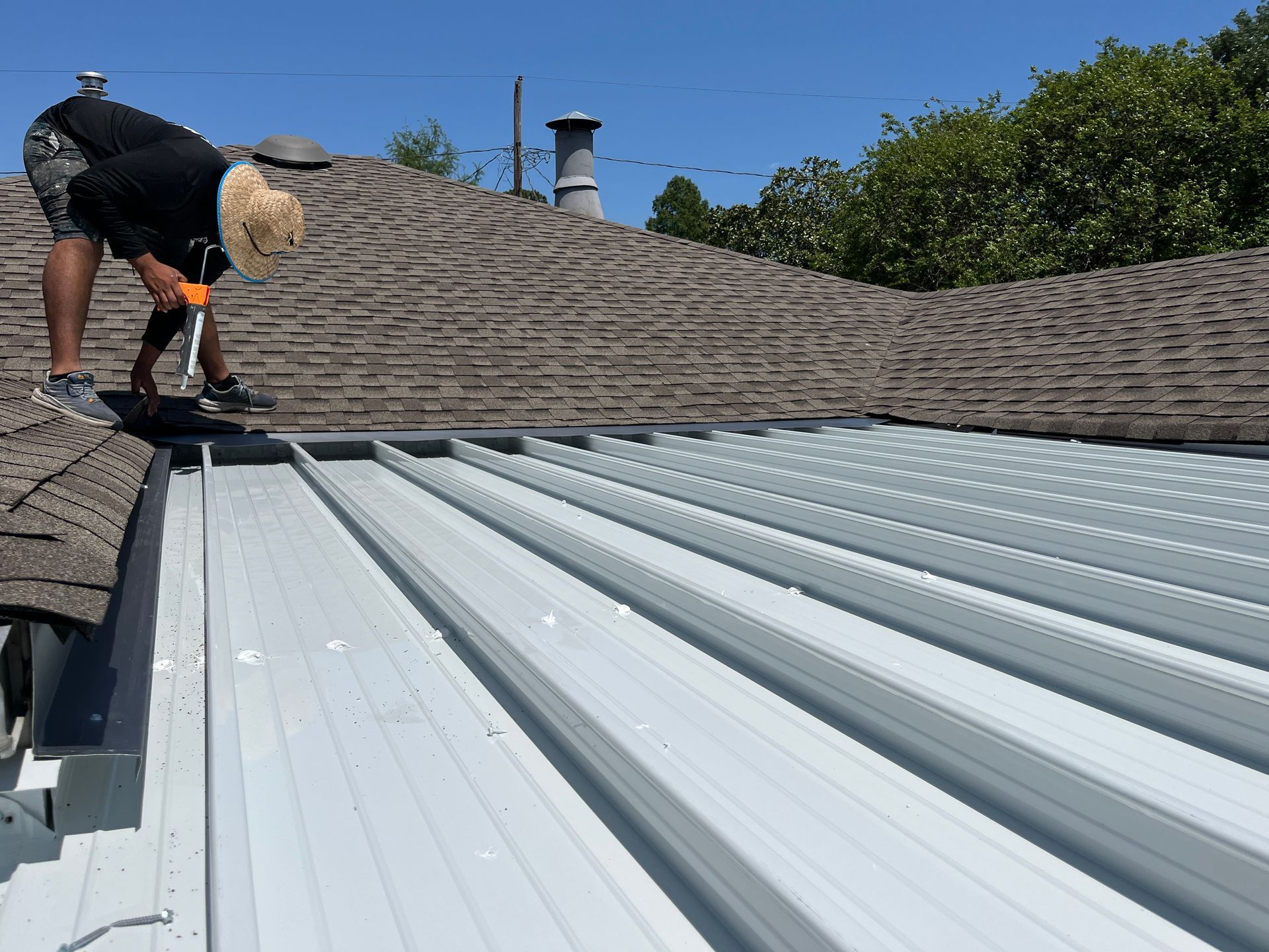 A worker wearing a straw hat and orange gloves installs metal roofing panels next to an existing shingled roof.