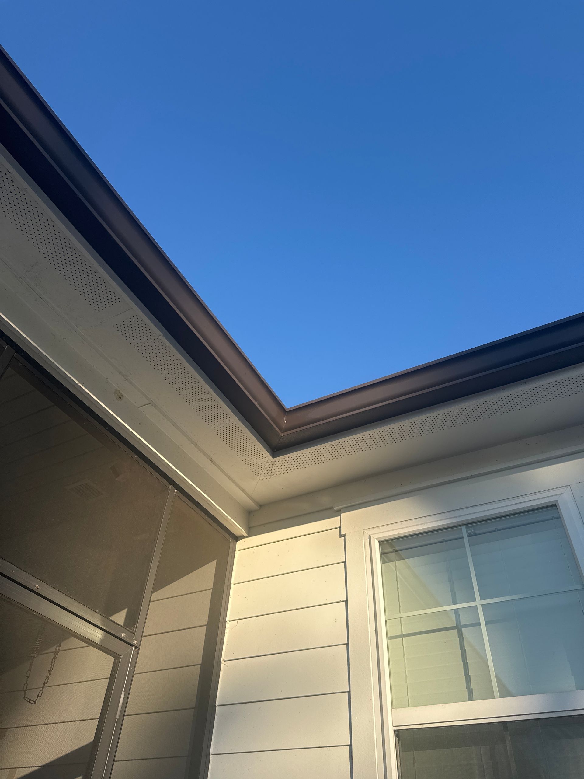 The corner of a home exterior showing white siding, a window, a screen enclosure, and dark roof eaves against a blue sky.