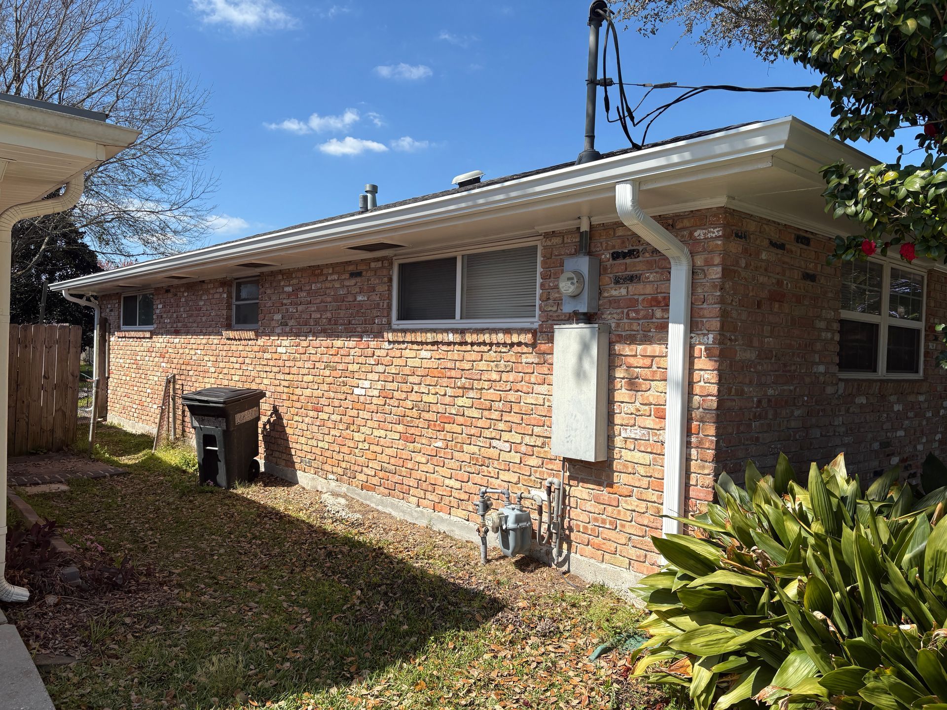 Exterior view of a single-story brick house with a white gutter, power meter, and a trash bin on a sunny day.