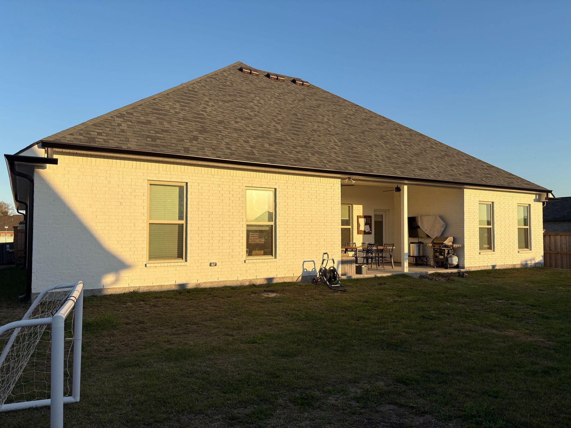 A rear view of a cream brick house with a dark roof and a covered patio, set against a clear blue sky at sunset.