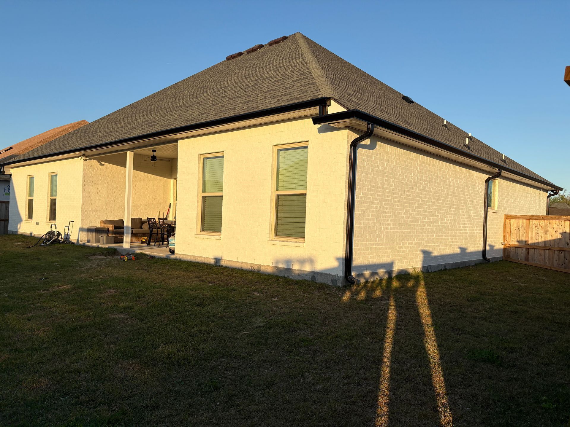 A beige brick house with a covered patio and a dark shingled roof under a clear blue sky.