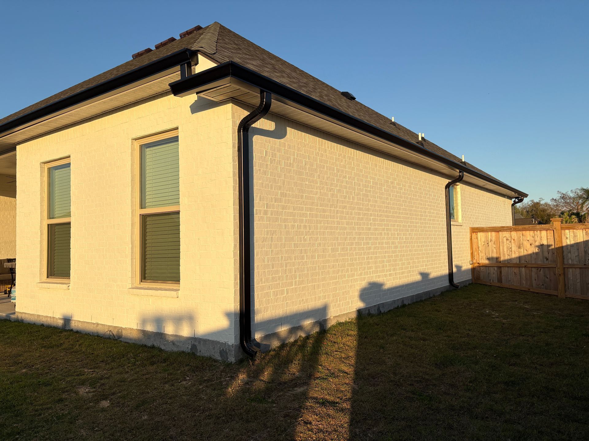 Side view of a light-colored brick house exterior with dark gutters, two windows, and a wood fence at sunset.