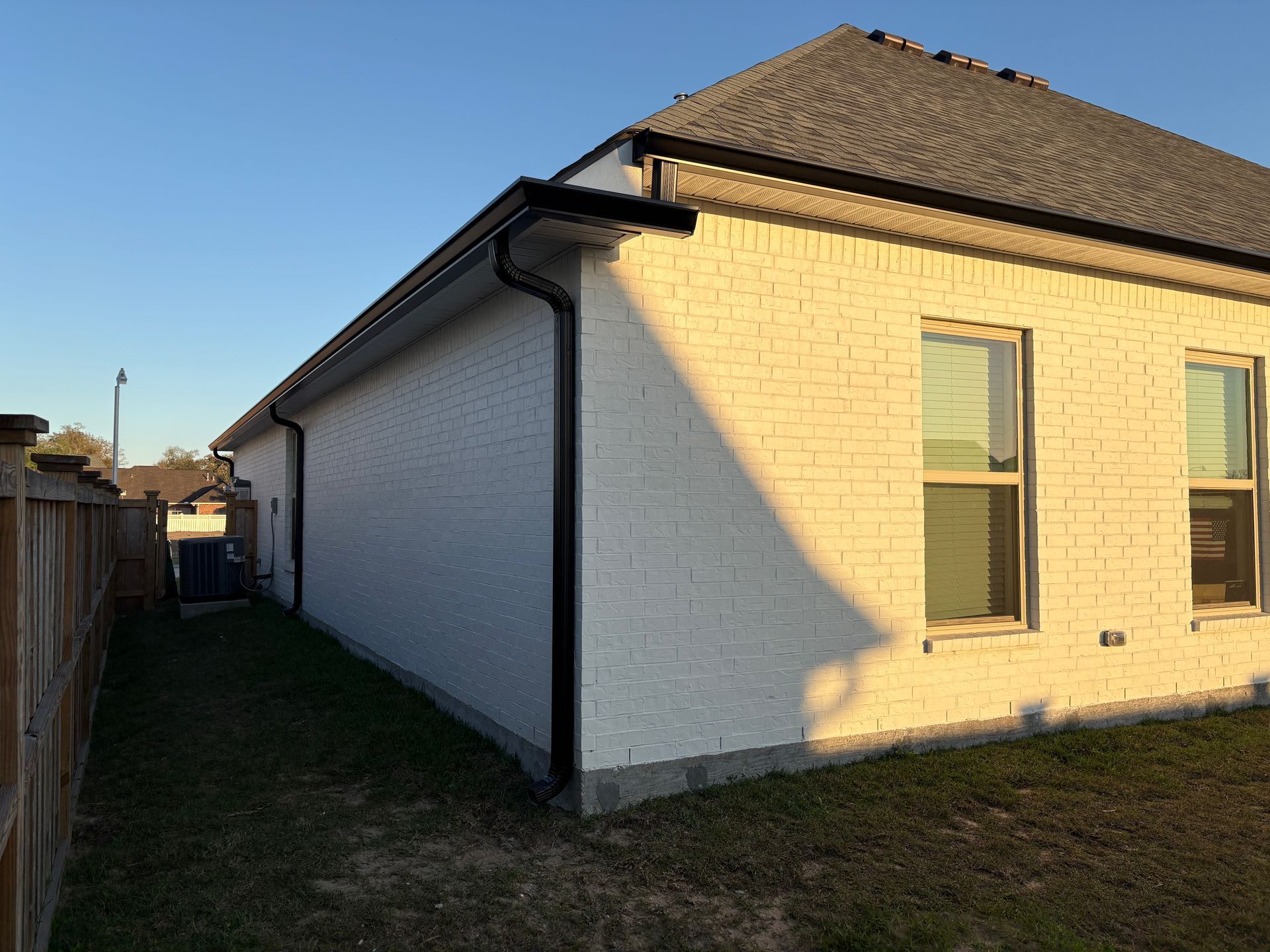 The side of a white brick house at sunset, featuring black gutters, two windows, and a neighboring wooden fence.