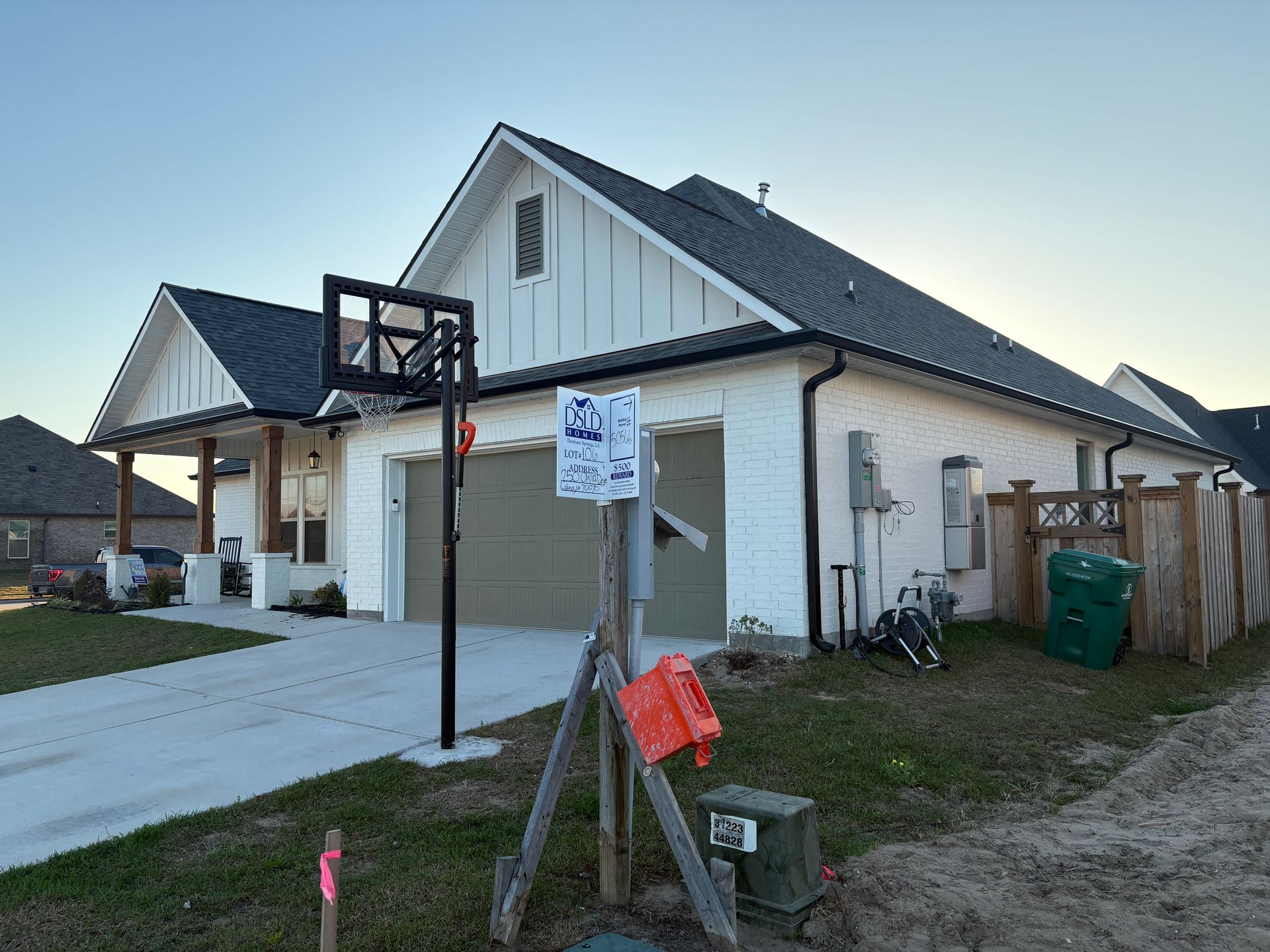 A white brick, modern farmhouse-style house with a gray roof, a basketball hoop, and a green trash bin in the yard.