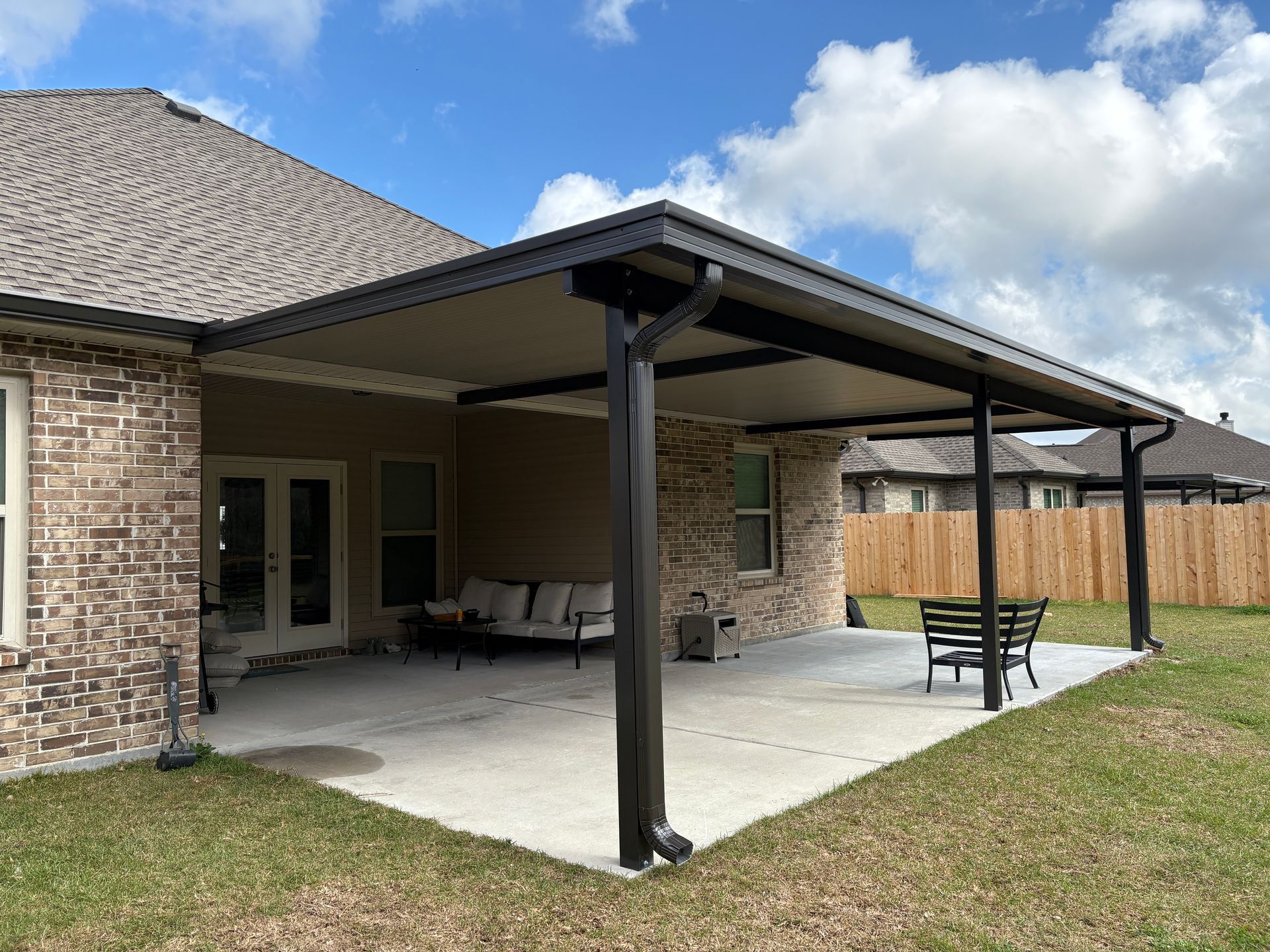 A brick house with a covered patio area featuring dark support beams and a concrete floor under a bright blue sky.