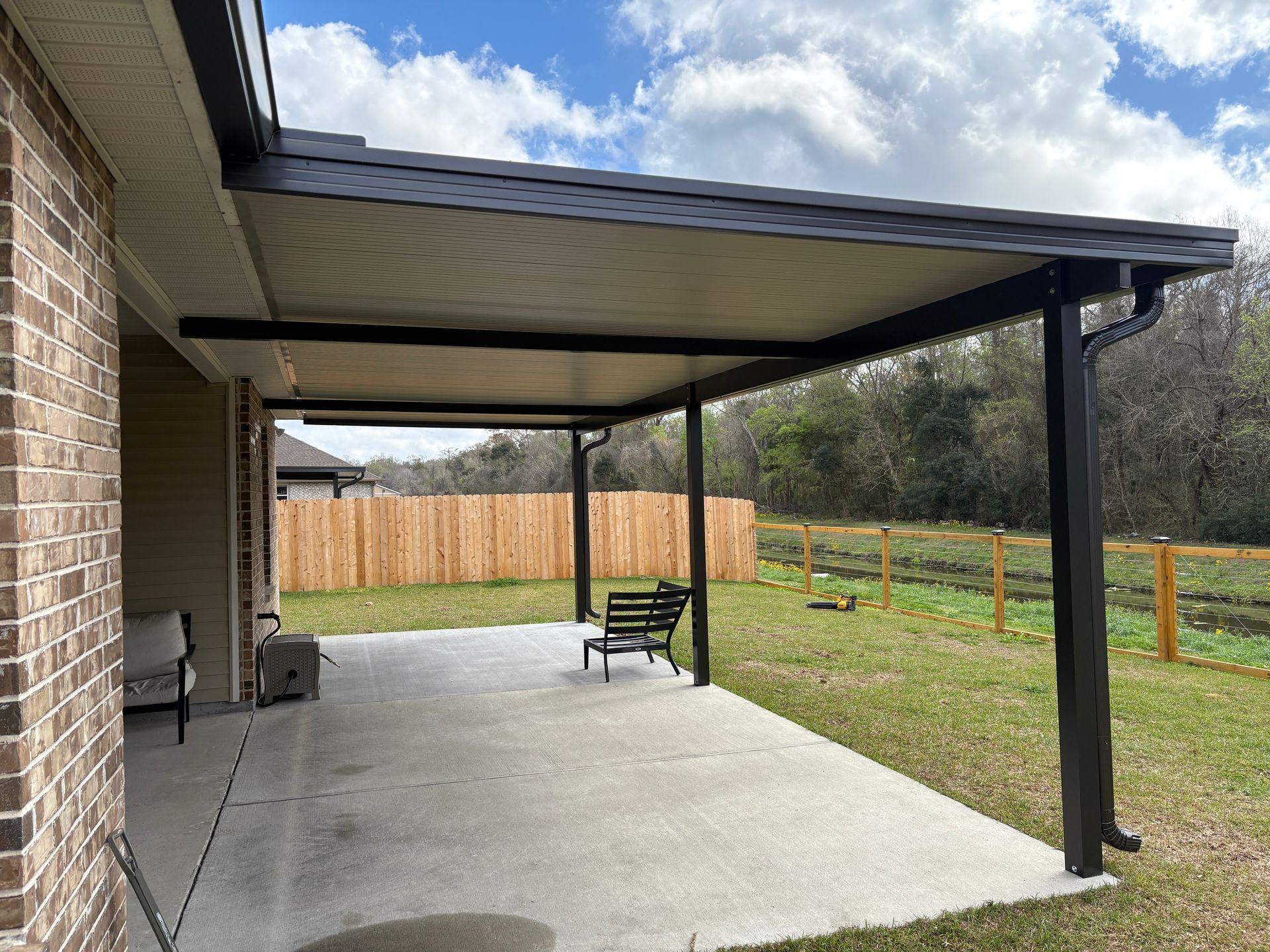 A backyard concrete patio with a dark, covered roof structure, a small bench, and a wooden fence in the background.