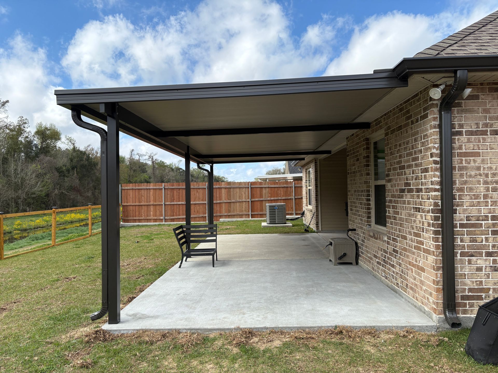 A covered concrete patio attached to a brick house, featuring a dark metal roof, support columns, and a bench.