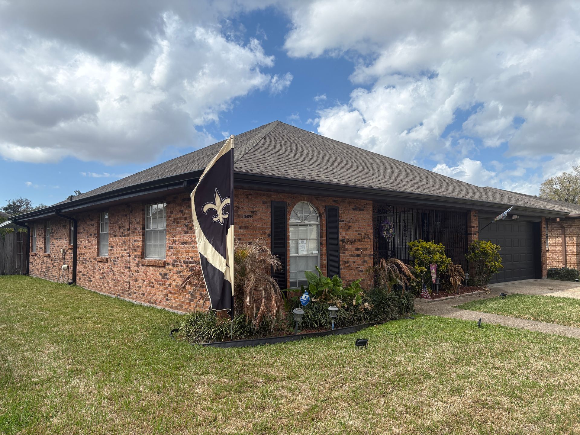 A single-story brick house with a dark roof and a New Orleans Saints flag on the front lawn under a cloudy blue sky.