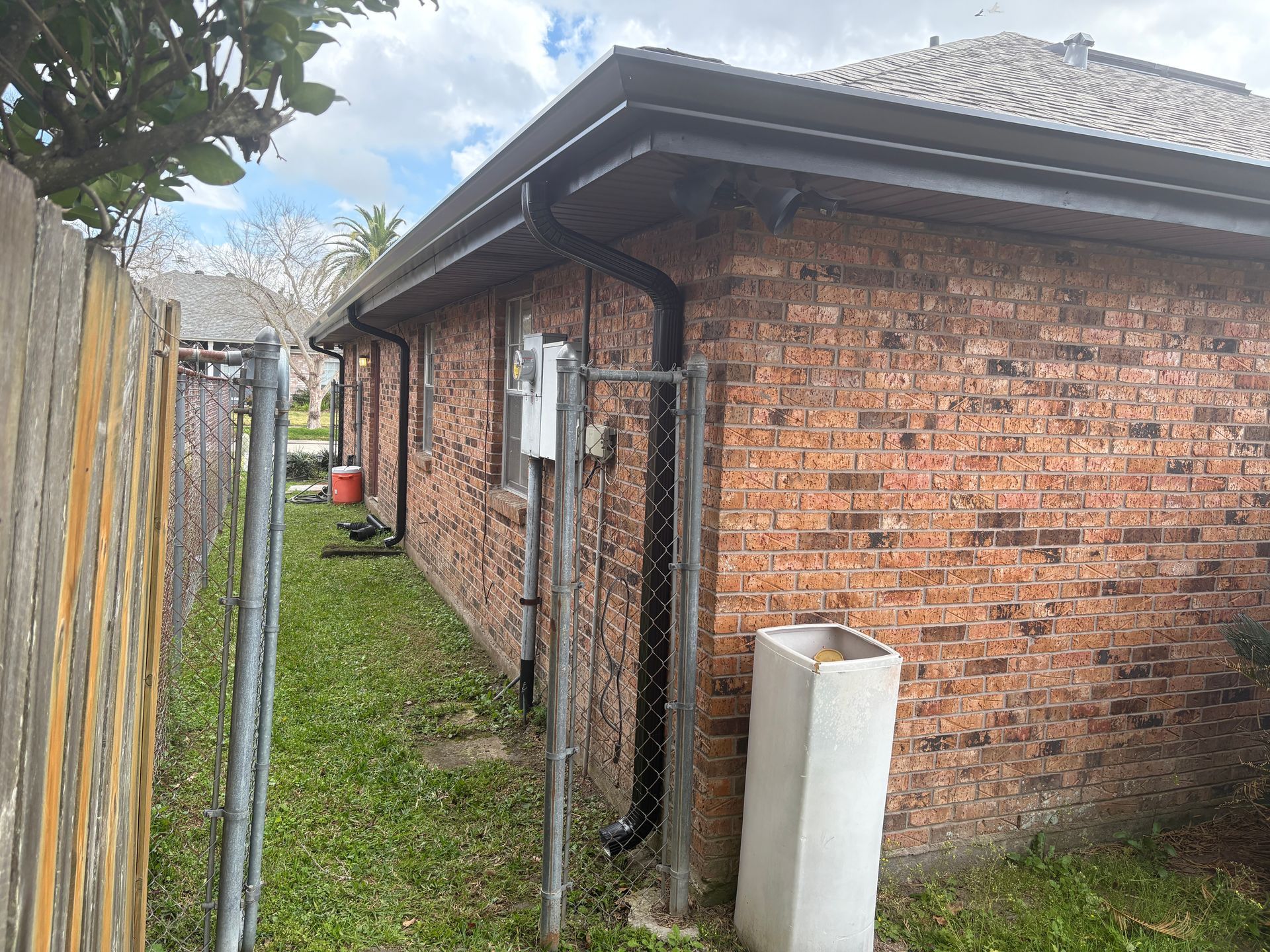 A side view of a brick house featuring dark gutters and downspouts, bordered by a chain-link fence in a grassy yard.