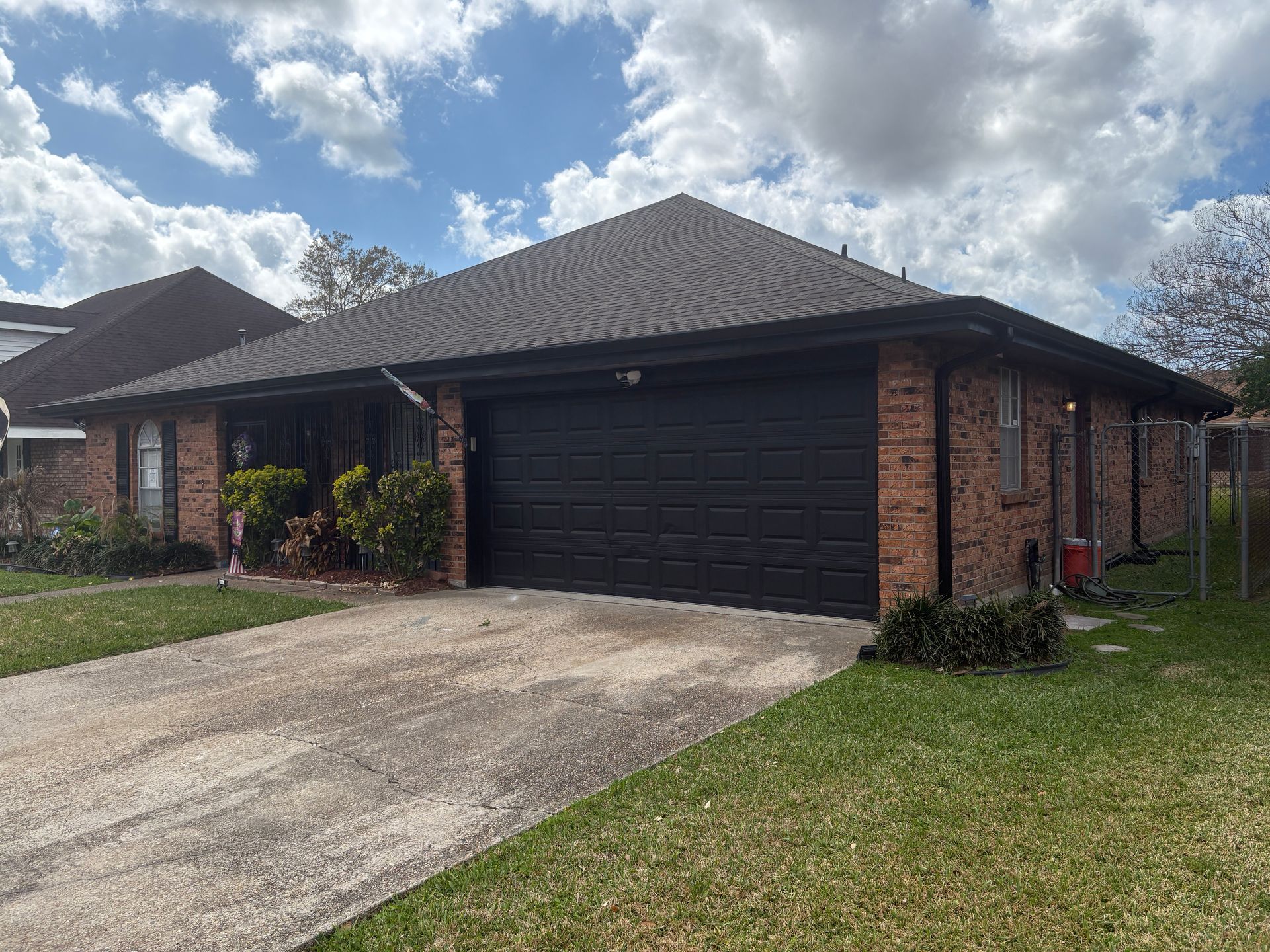 Single-story brick house with a dark roof and black two-car garage under a blue sky with scattered white clouds.