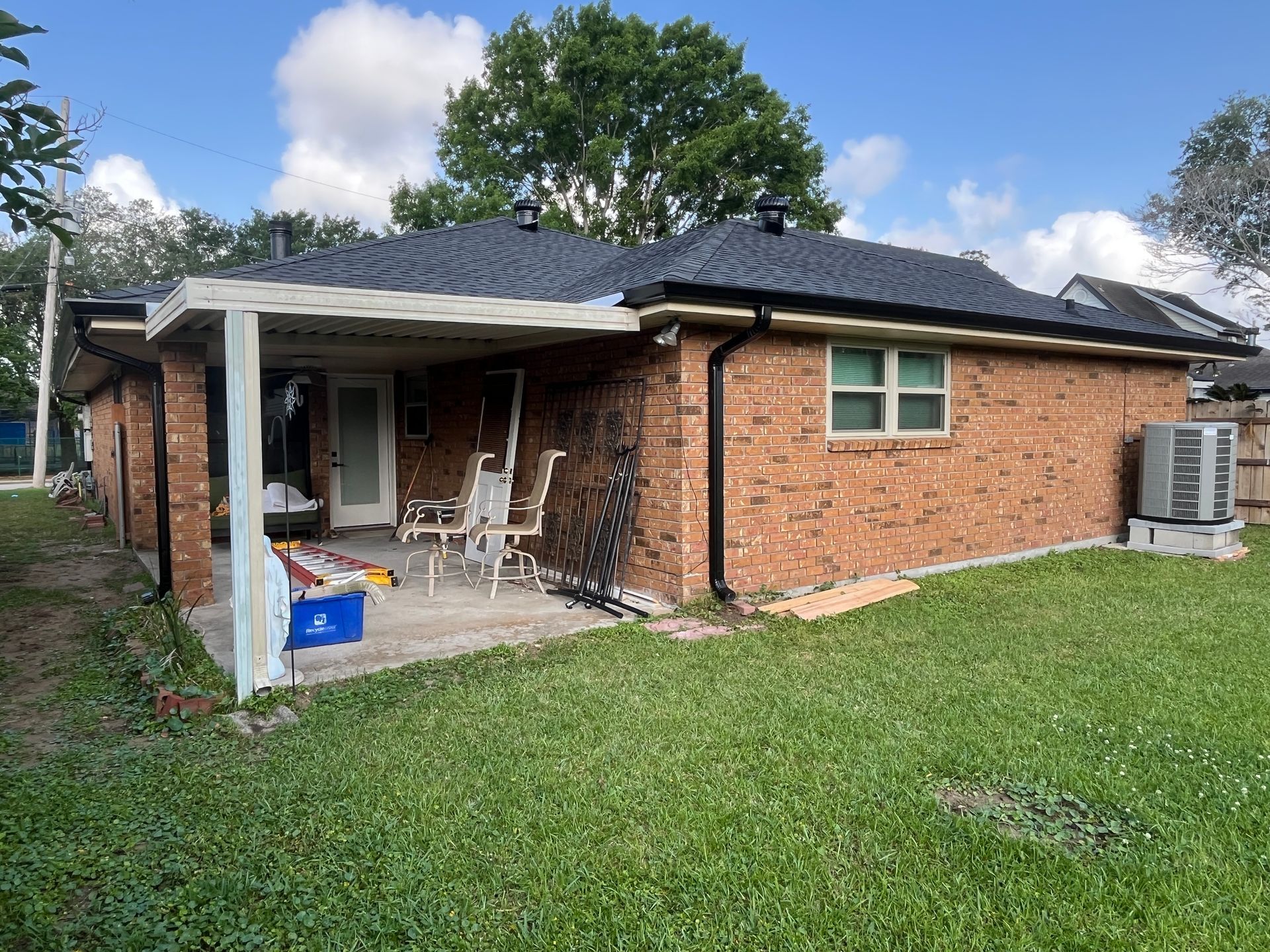 A brick house with a covered patio, black roof shingles, and a window, viewed from a grassy backyard under a sunny sky.
