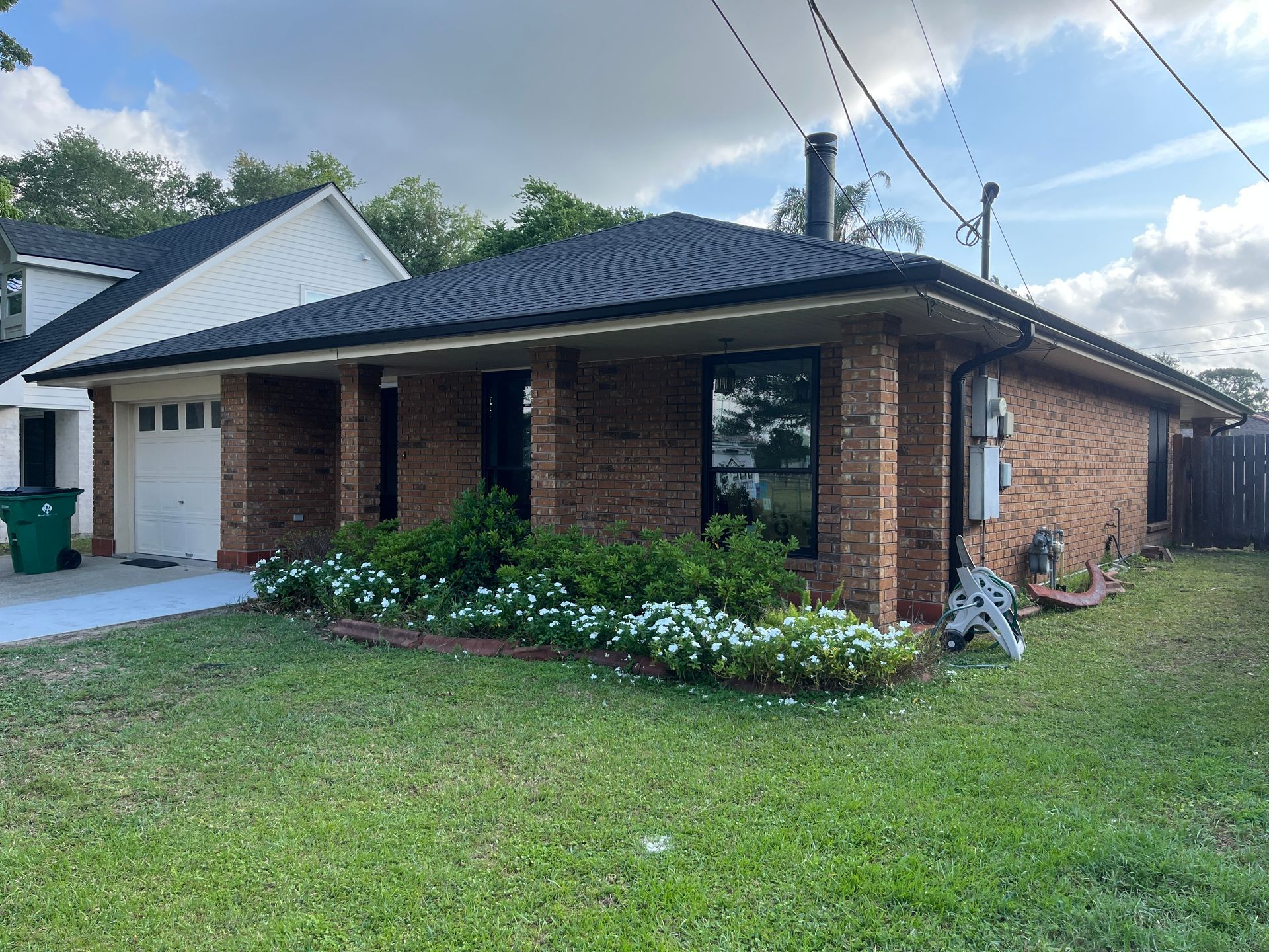 A brick one-story ranch house with a dark shingled roof, front porch pillars, and a patch of white flowers in the lawn.