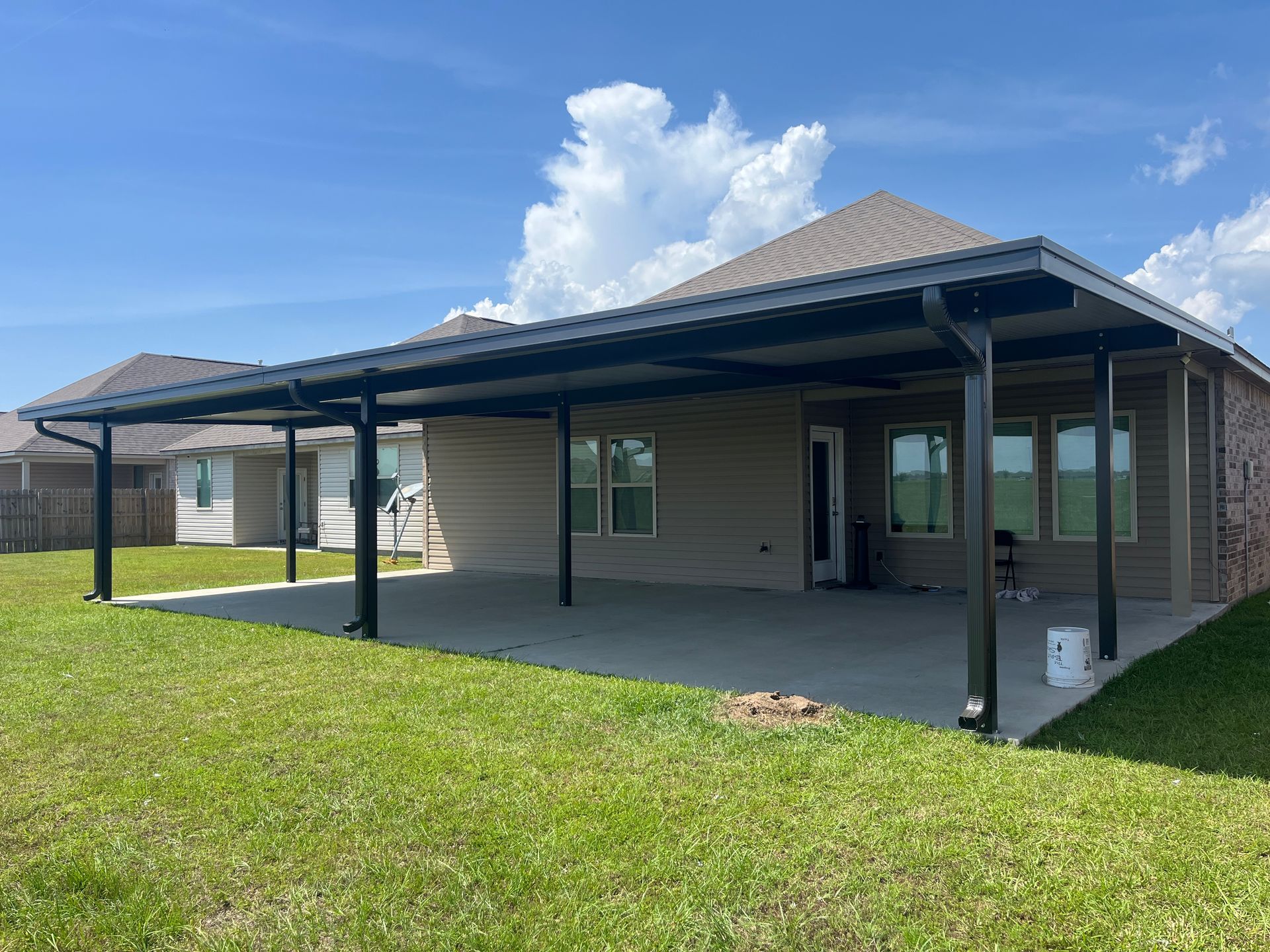 A tan house with a large black-framed patio cover extending over a concrete slab in a grassy backyard under a blue sky.