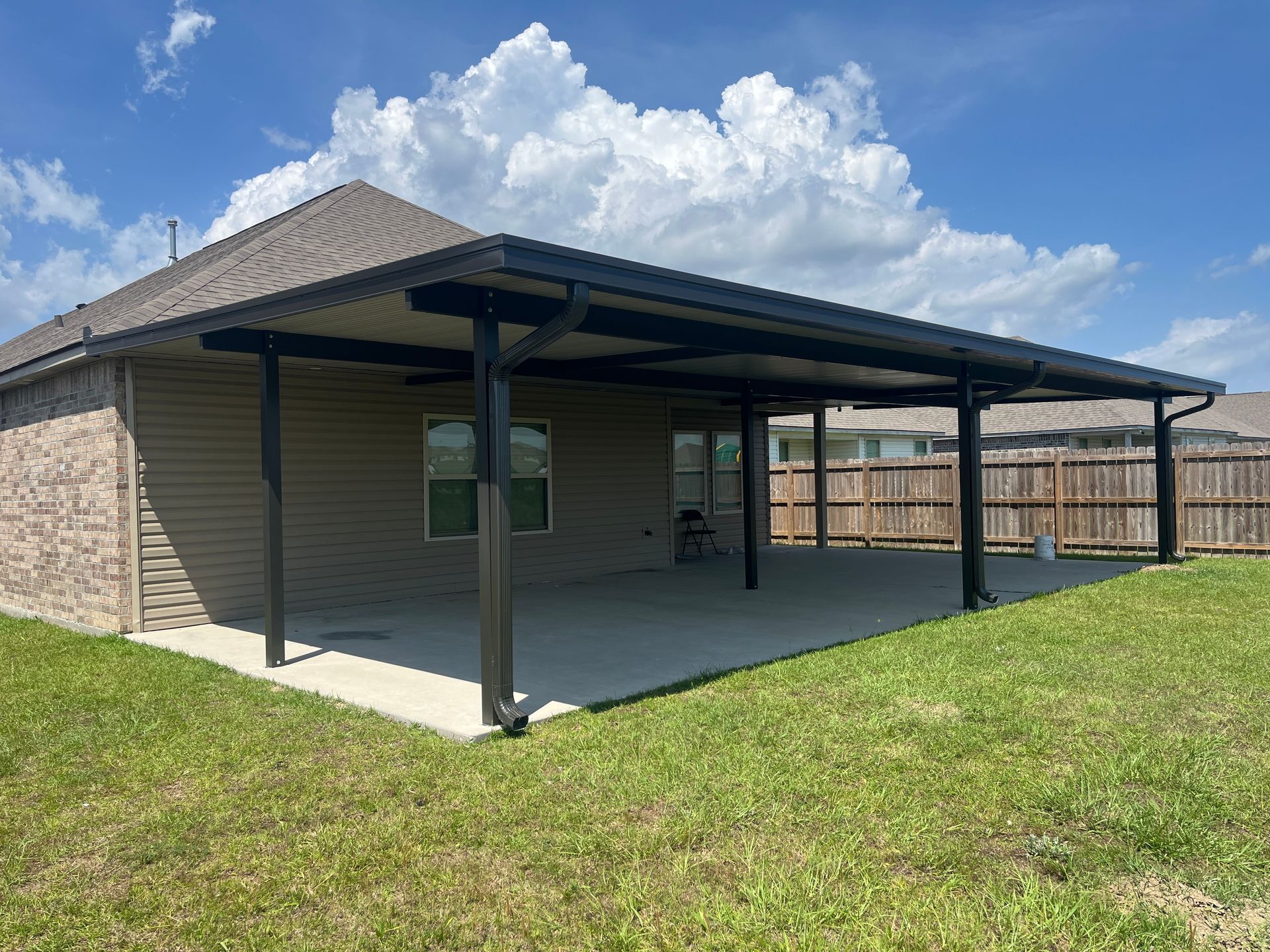A tan-brick house with a covered concrete patio, dark structural support beams, and a large backyard under a blue sky.