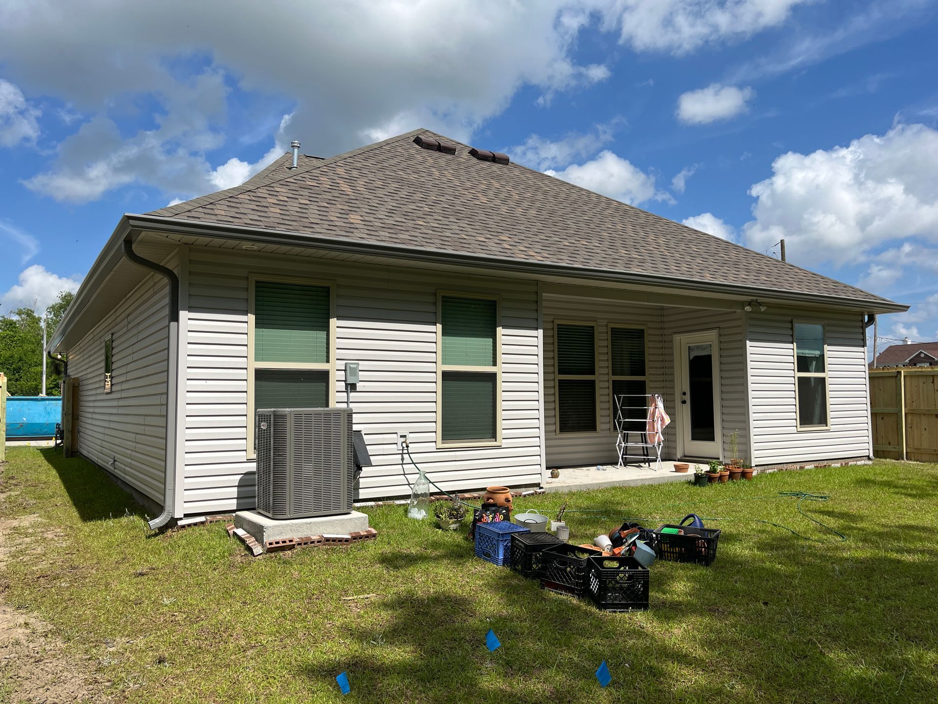 The back exterior of a tan vinyl-sided house with a shingled roof, a ground-level AC unit, and items scattered on the lawn.
