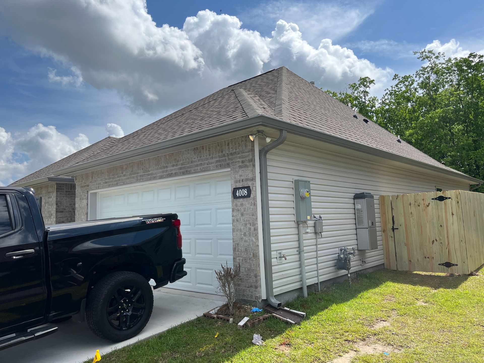A black pickup truck is parked in the driveway of a light brick and tan vinyl siding house with a tan shingle roof.