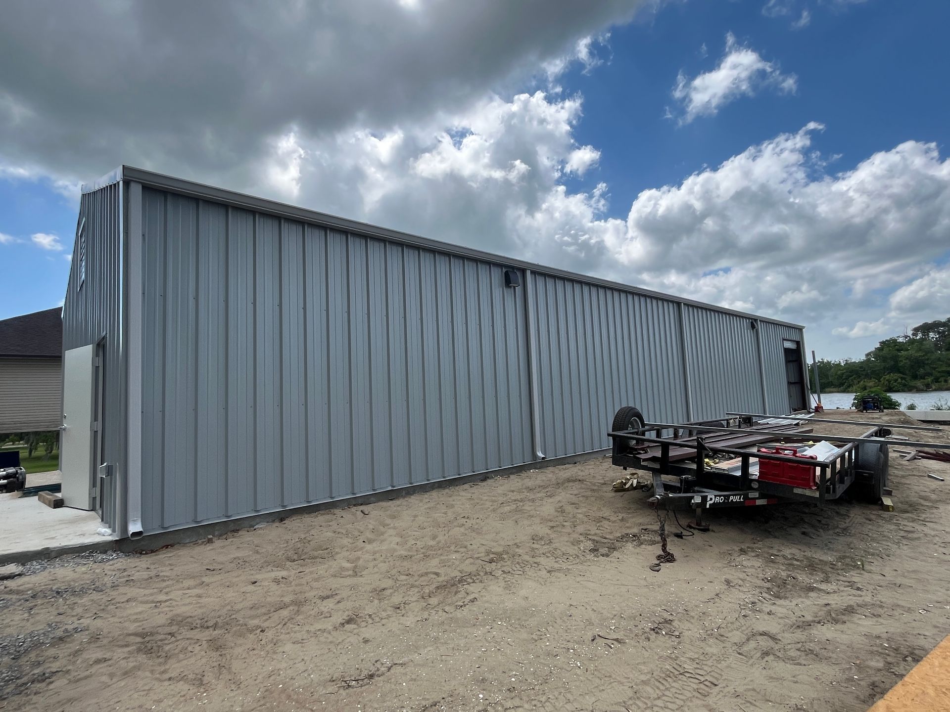 A large, light-gray metal building sits on a dirt lot with a trailer parked in front under a cloudy, blue sky.