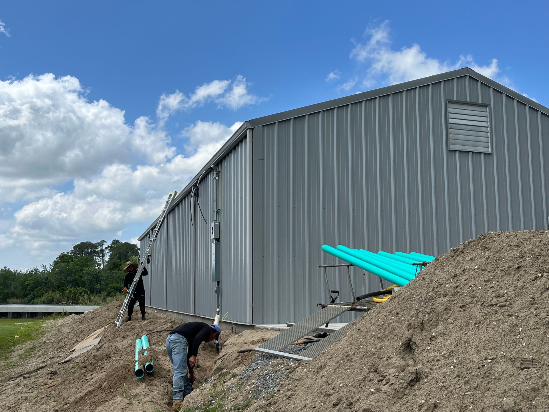 Two people work on the exterior of a gray metal building on a dirt slope under a blue sky with scattered clouds.