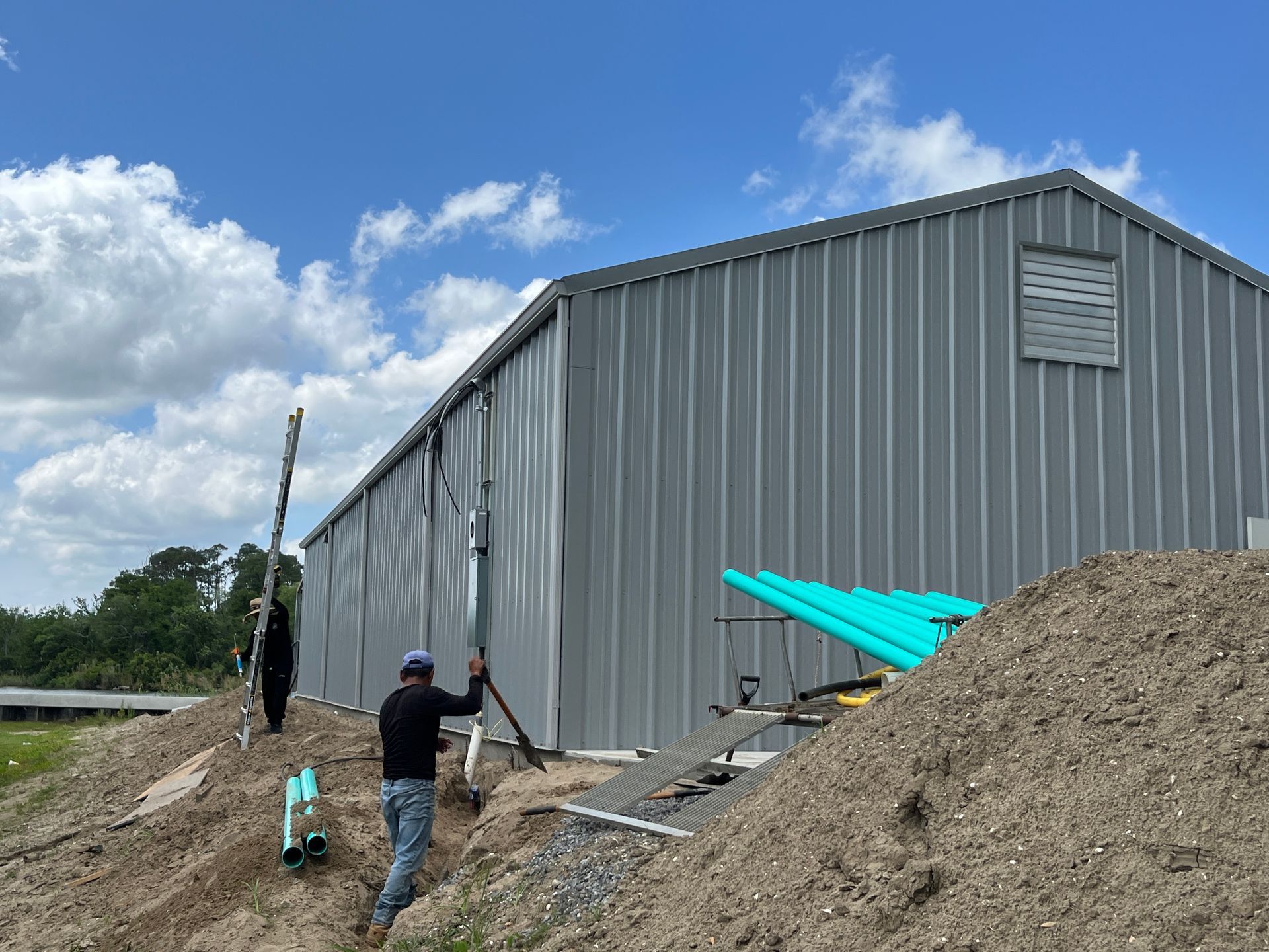 Two people work on a metal-sided building set on a dirt mound under a blue sky with clouds.